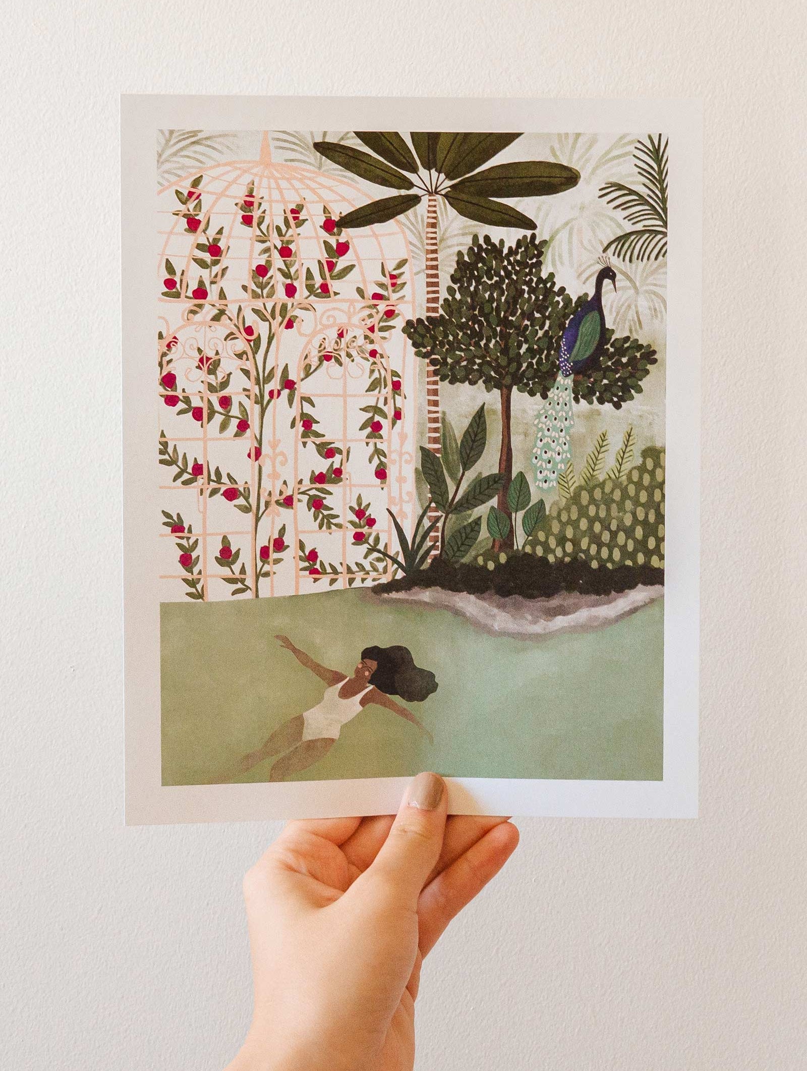 A hand holding up an illustration of a woman in the water, featuring the Peacock Garden illustration by mimi and august.