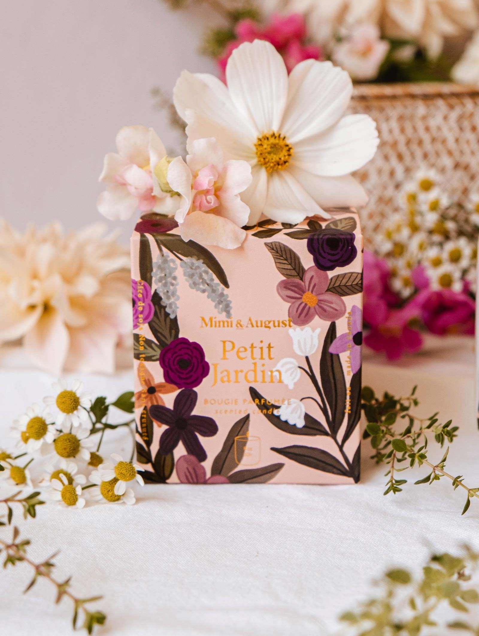 A floral-themed box labeled "Petit Jardin - Reusable Candle" from Mimi & August is set among fresh flowers in shades of white and pink, exuding a serene scent against a soft-focus background.