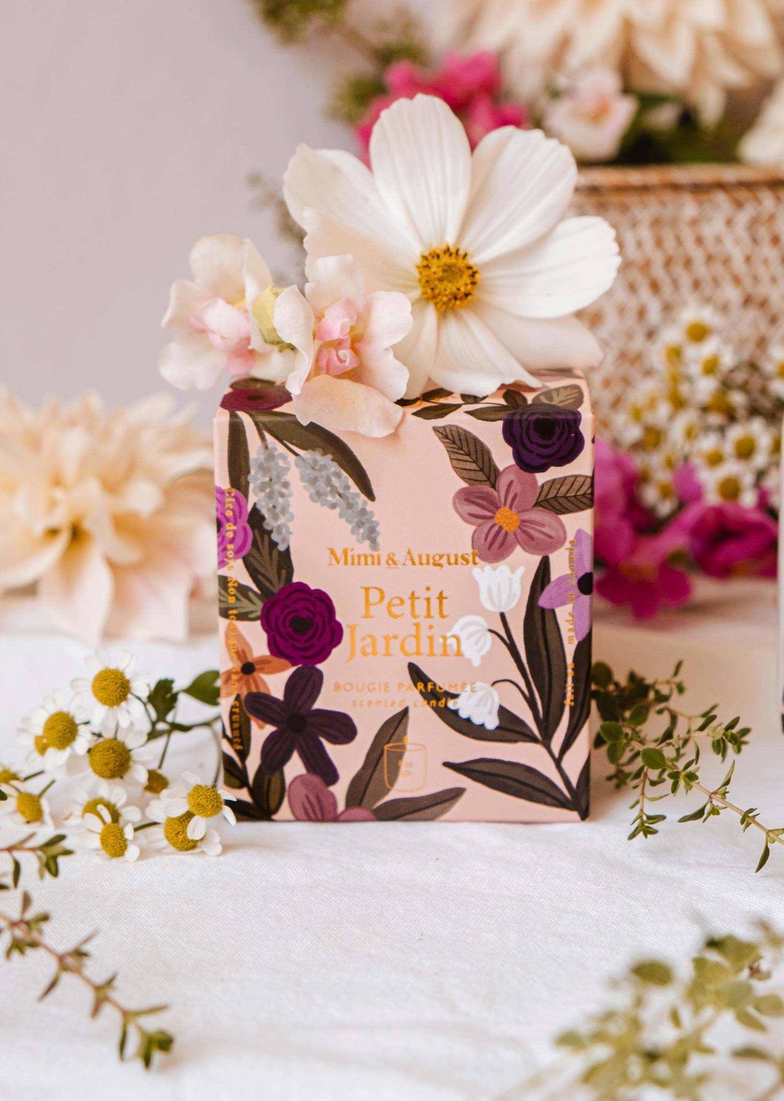Une boîte au thème floral étiquetée « Petit Jardin – Bougie réutilisable » de Mimi & August est disposée parmi des fleurs fraîches dans des tons de blanc et rose, dégageant un parfum serein sur un arrière-plan en flou doux.