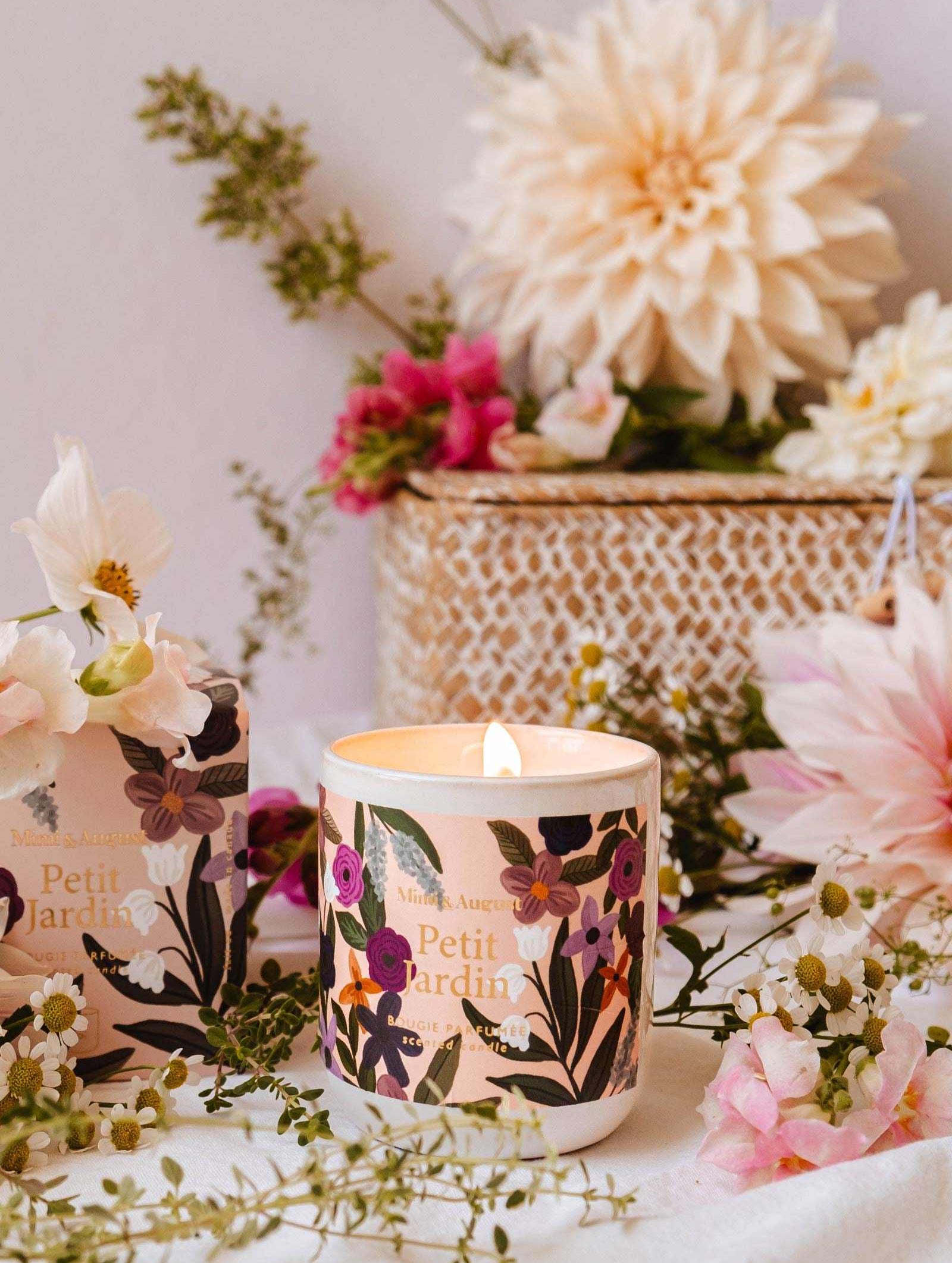 A Mimi & August Petit Jardin - Reusable Candle in a floral-themed jar sits on a table surrounded by small flowers and greenery, emanating a serene scent. In the background, there is a woven basket filled with white and pink fresh flowers from Petit Jardin.