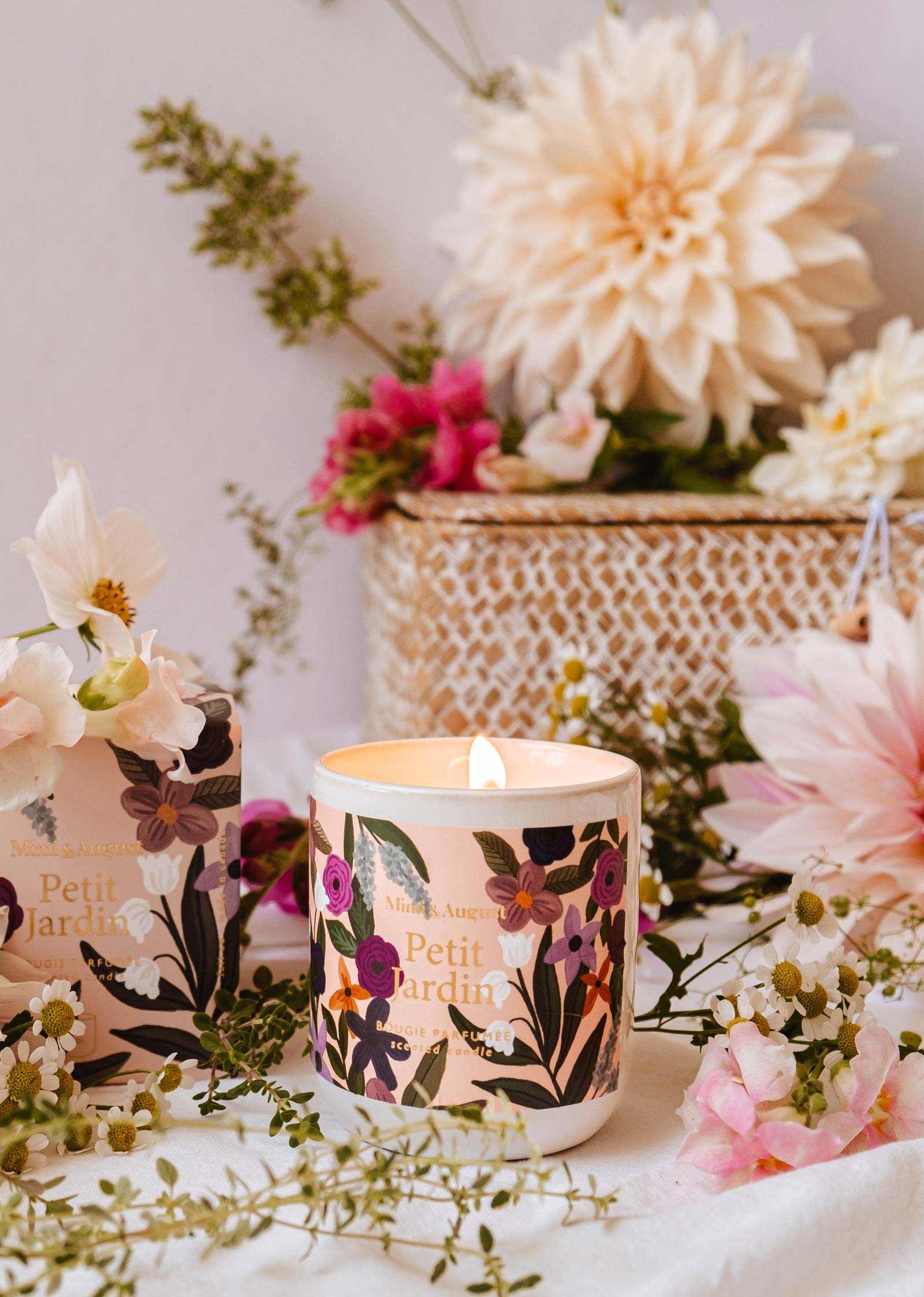Une bougie réutilisable Petit Jardin de Mimi & August dans un pot au thème floral repose sur une table entourée de petites fleurs et de verdure, émanant un parfum serein. En arrière-plan, un panier tressé rempli de fleurs fraîches blanches et roses.