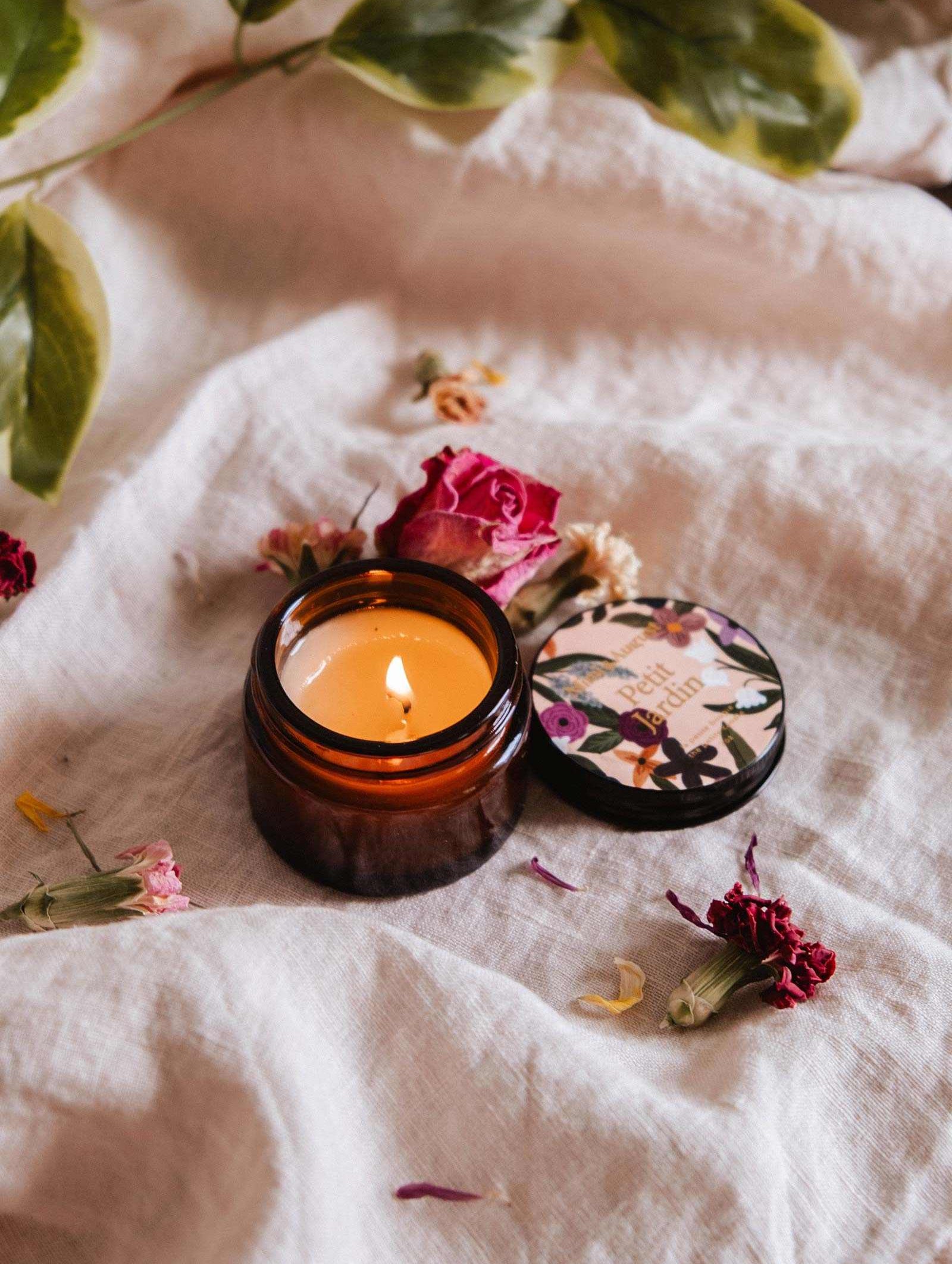 La bougie de soya Petit Jardin 2 oz de Mimi & August est une bougie réutilisable coulée à la main dans un petit pot floral. Ouverte et allumée, elle repose sur un tissu blanc avec des fleurs séchées et des feuilles vertes, ajoutant du charme à toute pièce. Faite à Montréal.