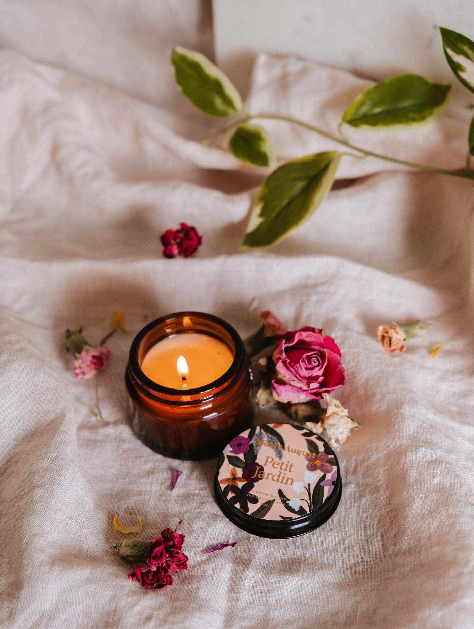 La bougie de soya Petit Jardin 2 oz de Mimi & August, au parfum floral, est coulée à la main à Montréal et repose sur un tissu clair avec un couvercle décoratif en étain et des fleurs séchées et feuilles éparpillées.
