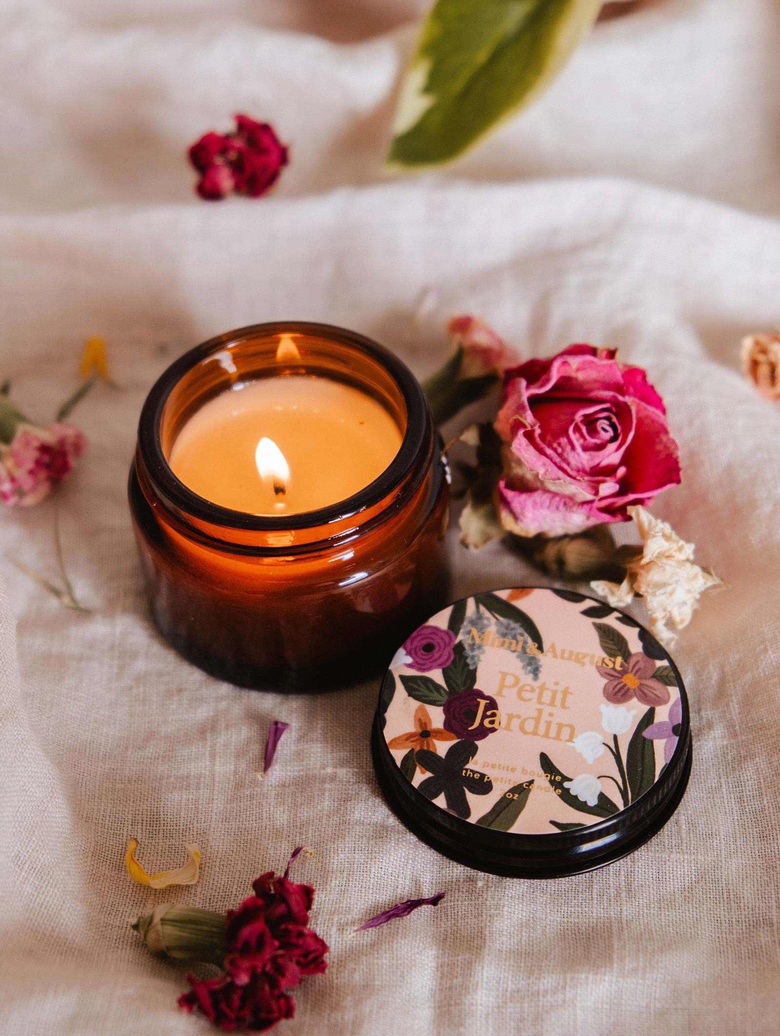 Une bougie de soya Petit Jardin 2 oz de Mimi & August allumée dans un pot en verre brun repose sur un tissu avec des fleurs séchées et un couvercle décoratif. Cette bougie réutilisable est coulée à la main à Montréal.