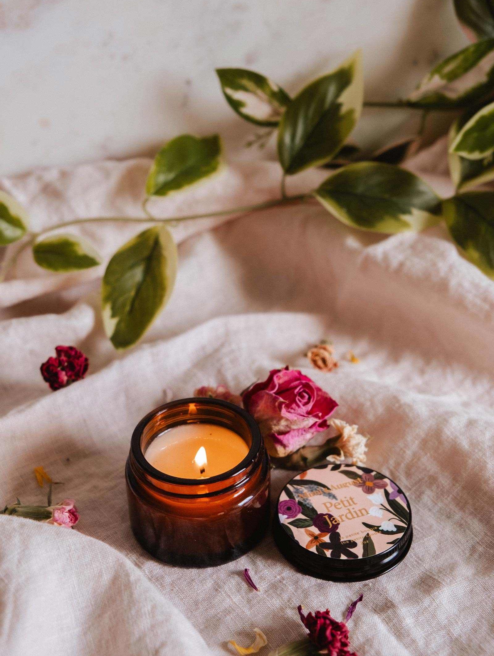 La bougie de soya Petit Jardin 2 oz de Mimi & August, dans un pot en verre brun avec son couvercle à proximité, repose sur un tissu beige entourée de fleurs séchées et de feuilles vertes.