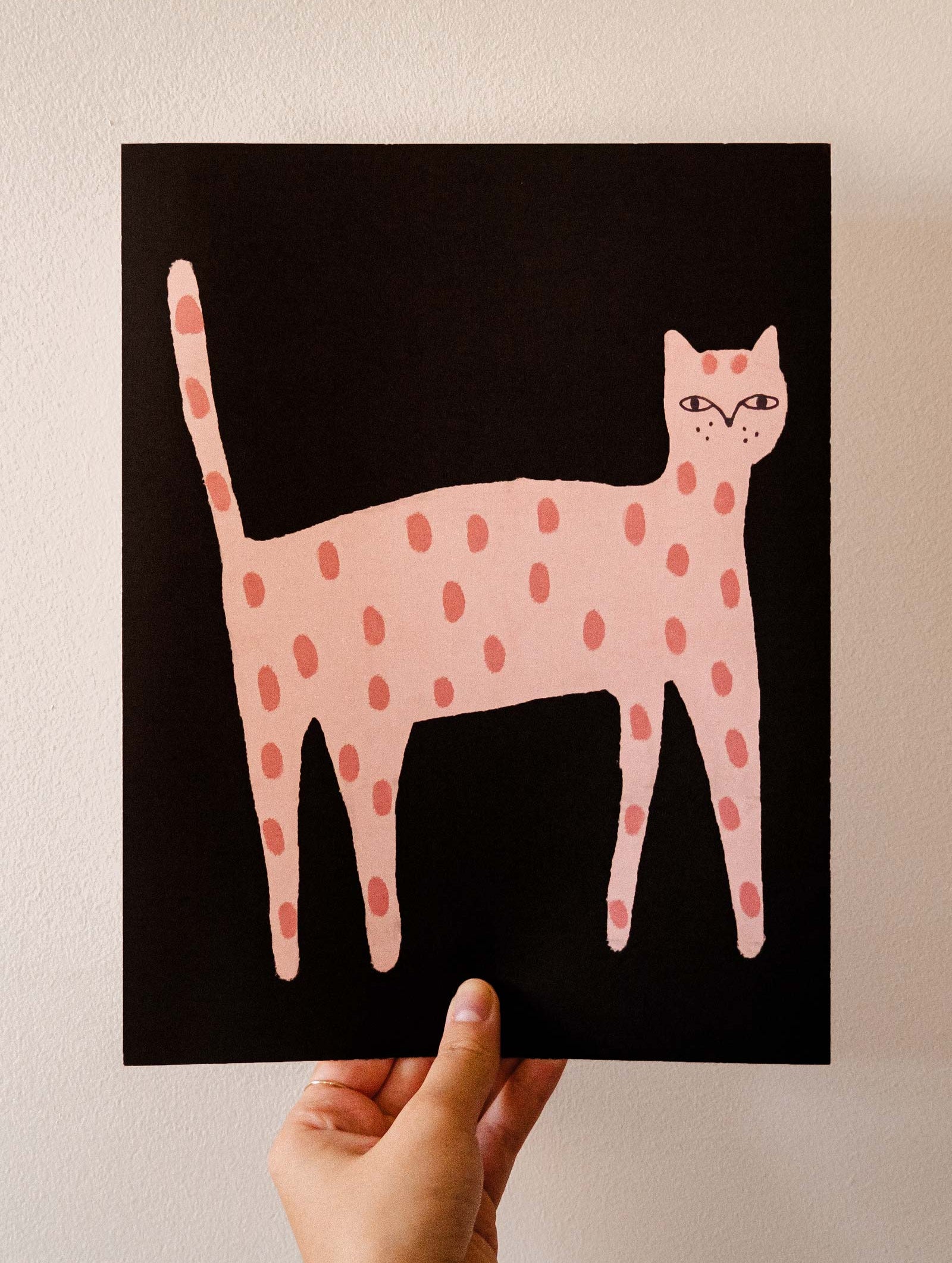 Hand holding a Playful Cat Art Print by Mimi & August featuring a stylized pink cat with light pink spots on a black background, all beautifully printed on recycled paper.