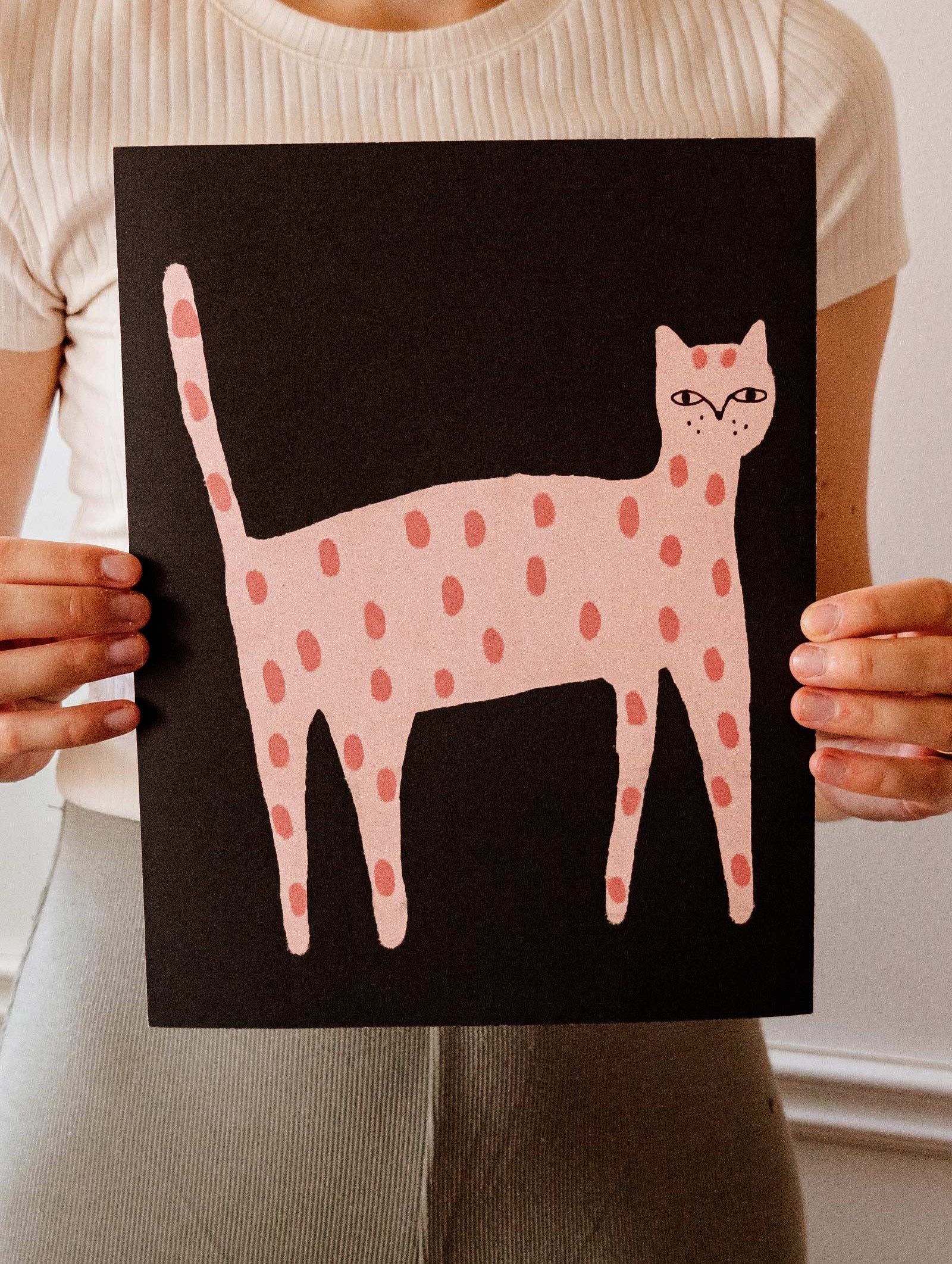 A person holds up a black poster featuring a pink, polka-dotted cat illustration. This Playful Cat Art Print by Mimi & August is crafted on recycled paper, adding an eco-friendly touch.