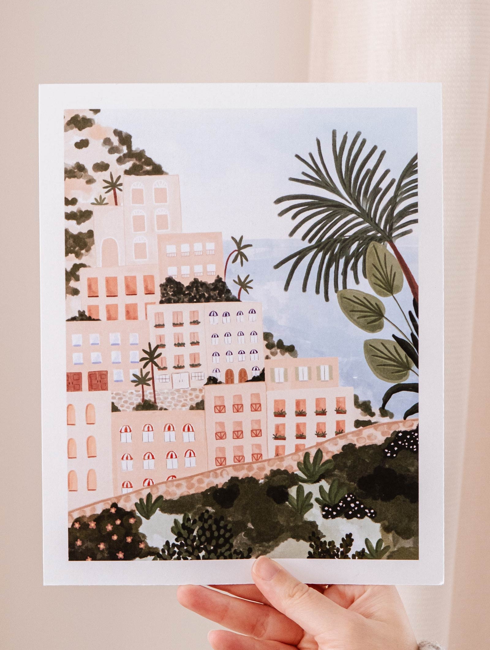 A hand holding up a Mimi & August Positano Art Print of Positano, an Italian coastal town with palm trees.