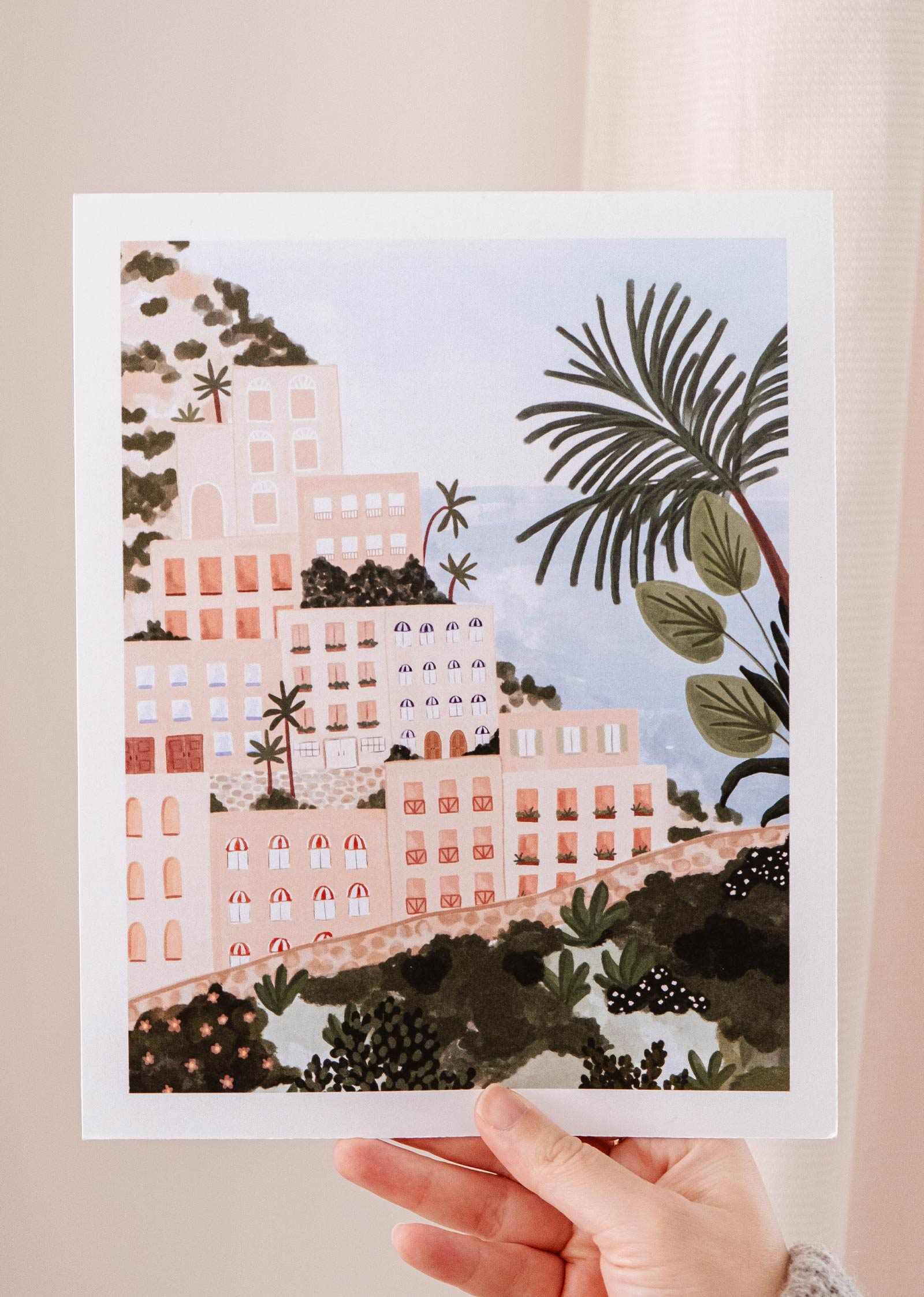 A hand holding up a Mimi & August Positano Art Print of Positano, an Italian coastal town with palm trees.