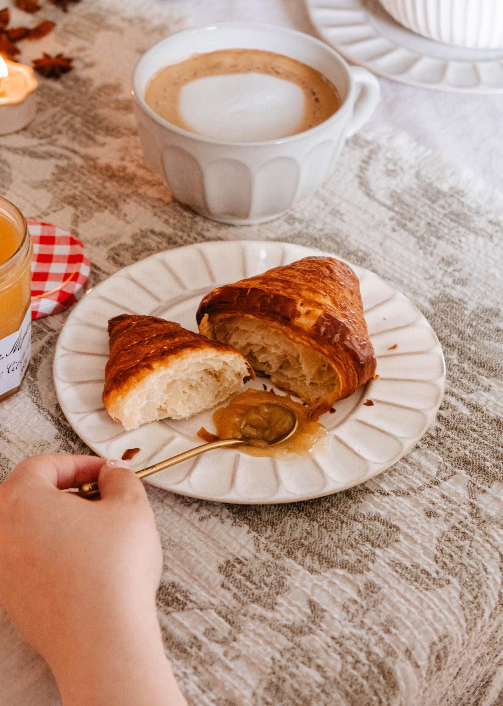 A hand holds a spoon with honey above handmade ceramic plate by Mimi & August, which displays a sliced croissant beside a cup of coffee on a table draped with a white tablecloth.