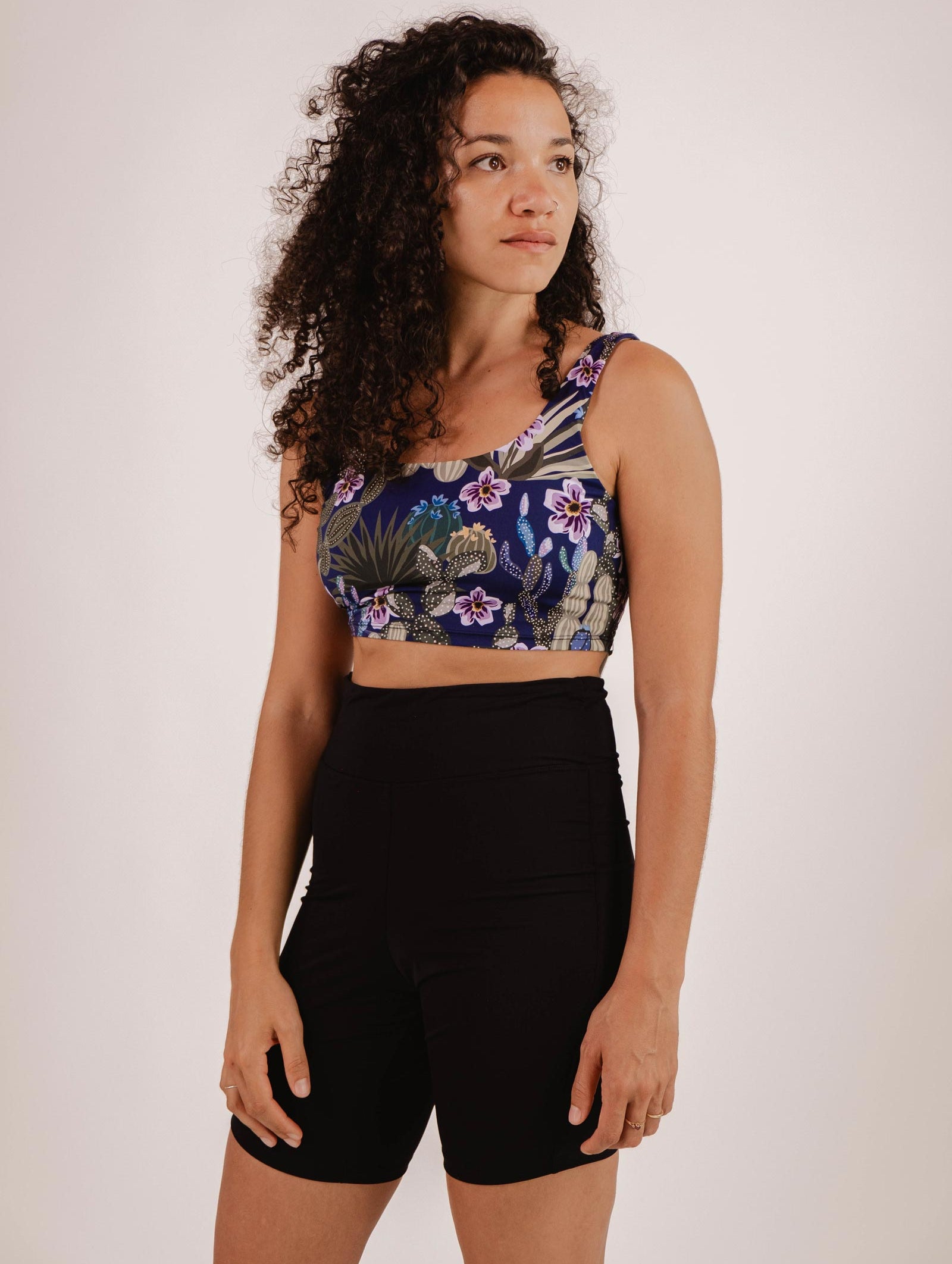 A person with curly hair is looking off-camera, wearing a floral crop top and black high-waisted **Riviera Black High Waist Swim Bike Short by Mimi & August** against a plain background.