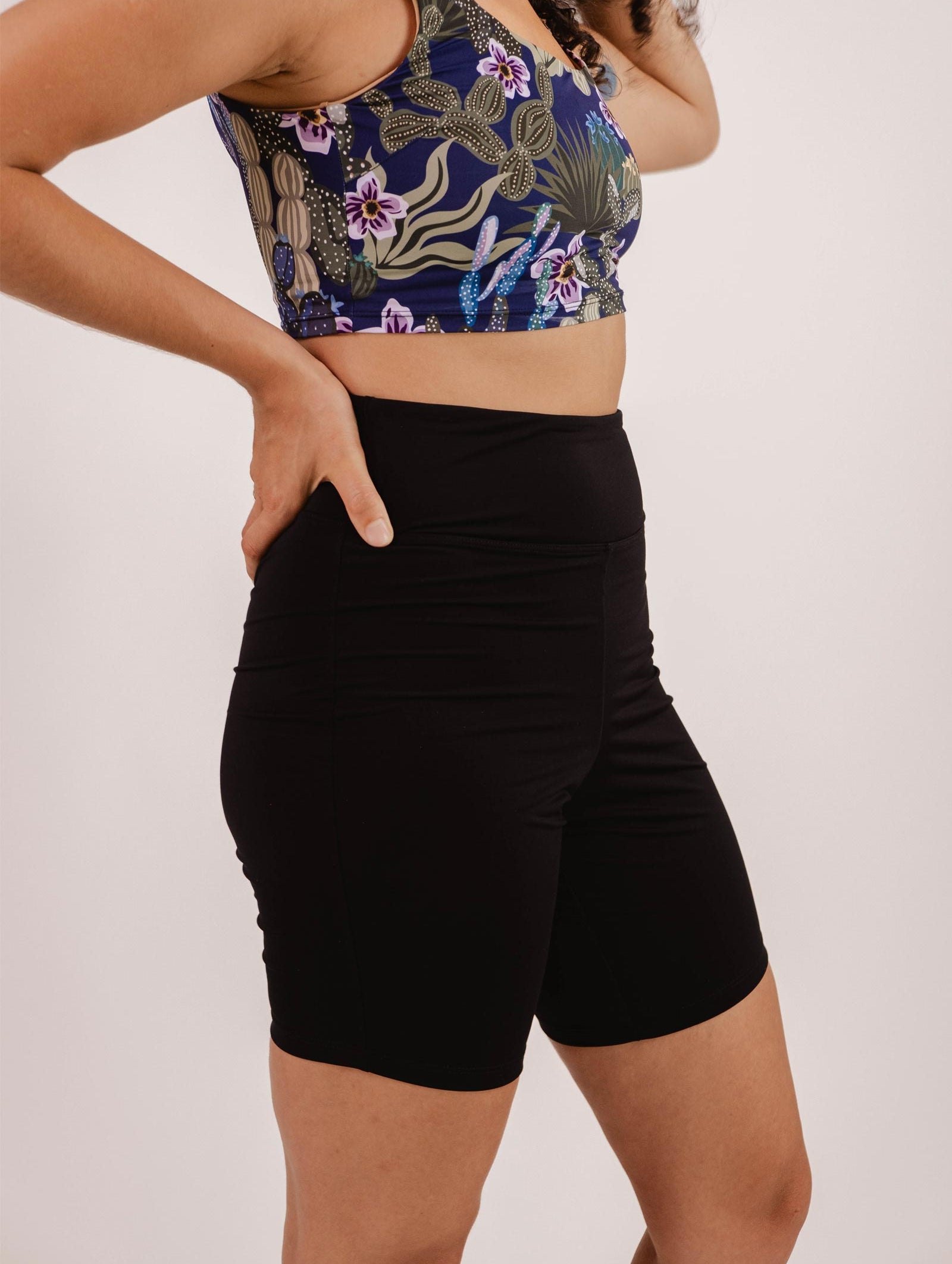 A person wearing a floral-patterned sports bra and Mimi & August's Riviera Black High Waist Swim Bike Short made from Econyl swimsuit fabric stands with hands on hips.