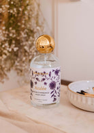 A bottle of Mimi & August's Azalée Room Spray with a floral design and gold cap sits on a table beside dried flowers, filling the air with jasmine and white tea notes.