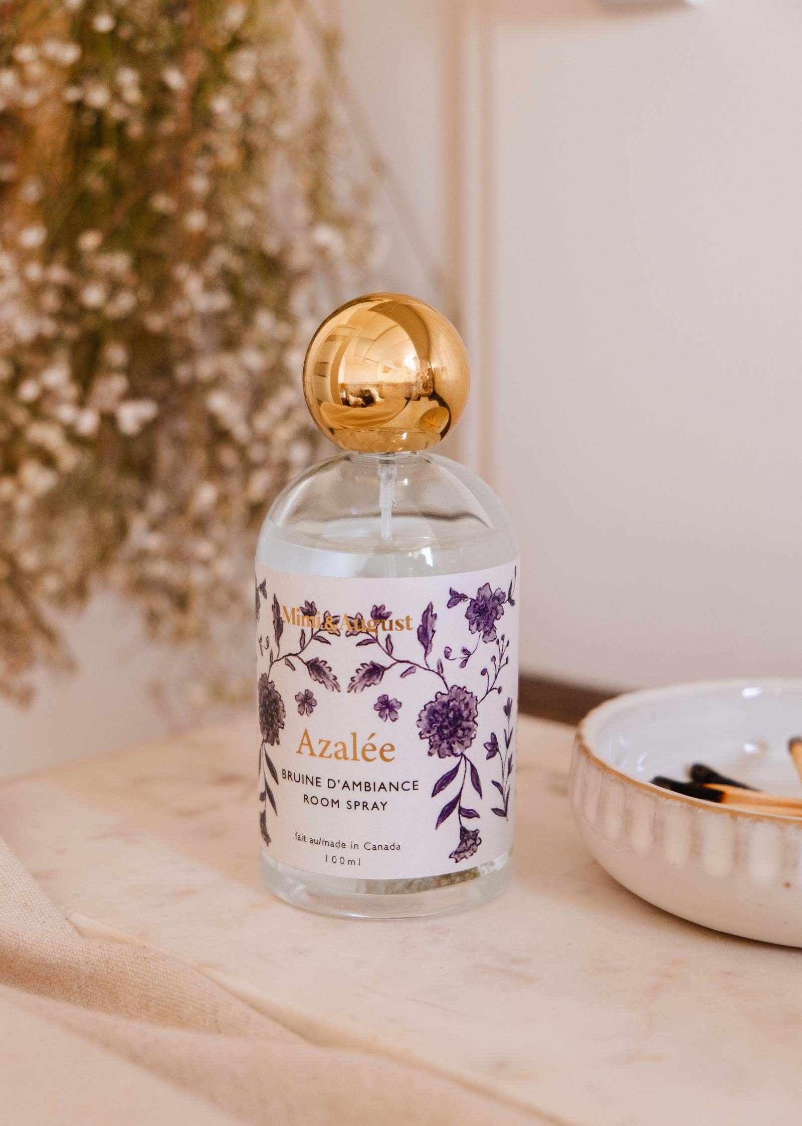 A bottle of Mimi & August's Azalée Room Spray with a floral design and gold cap sits on a table beside dried flowers, filling the air with jasmine and white tea notes.