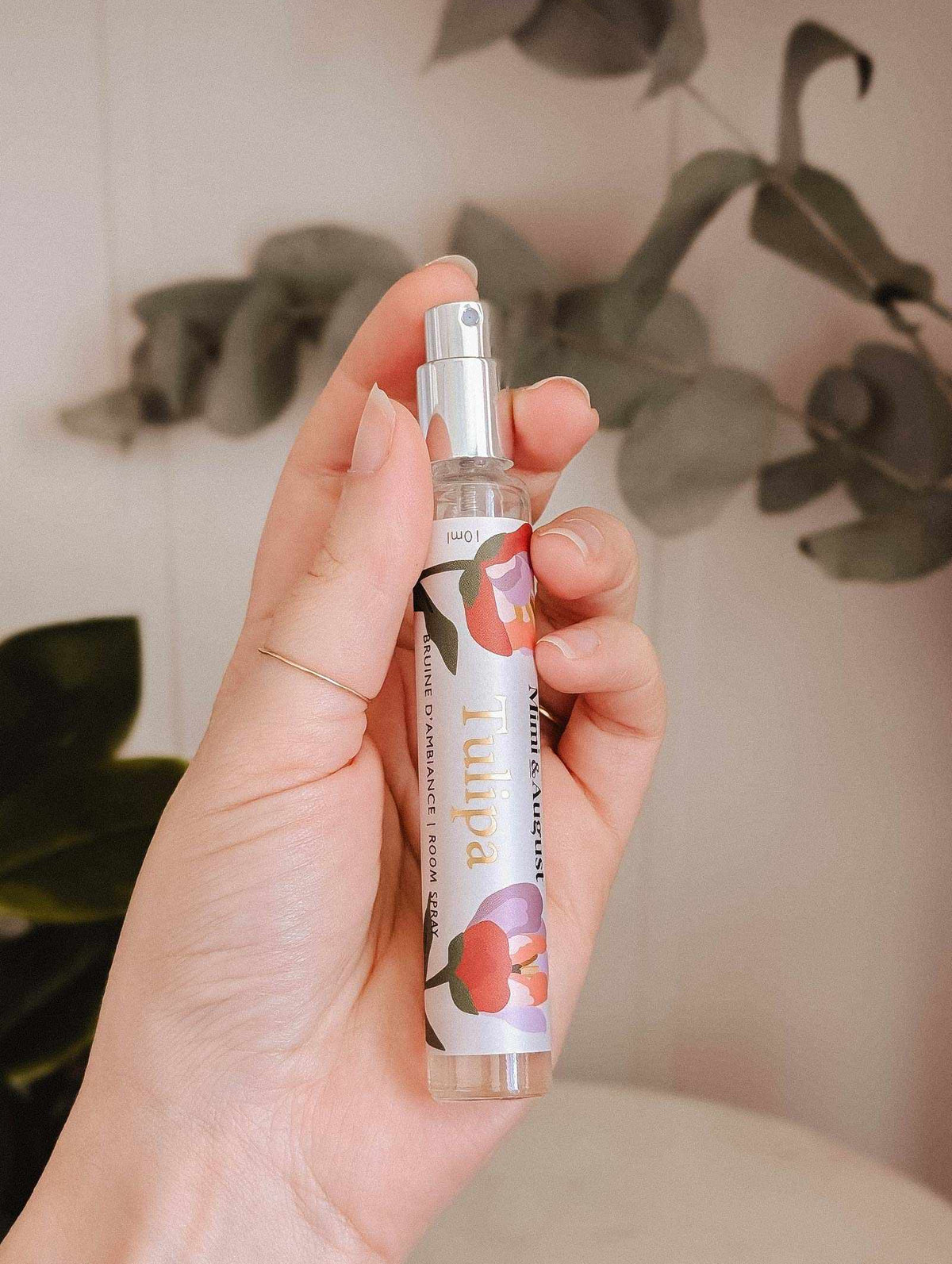 A hand holds a Mimi & August Room Spray Tulipa bottle, featuring a floral design, with a blurred leafy background that highlights the essence of this fragrant spray.