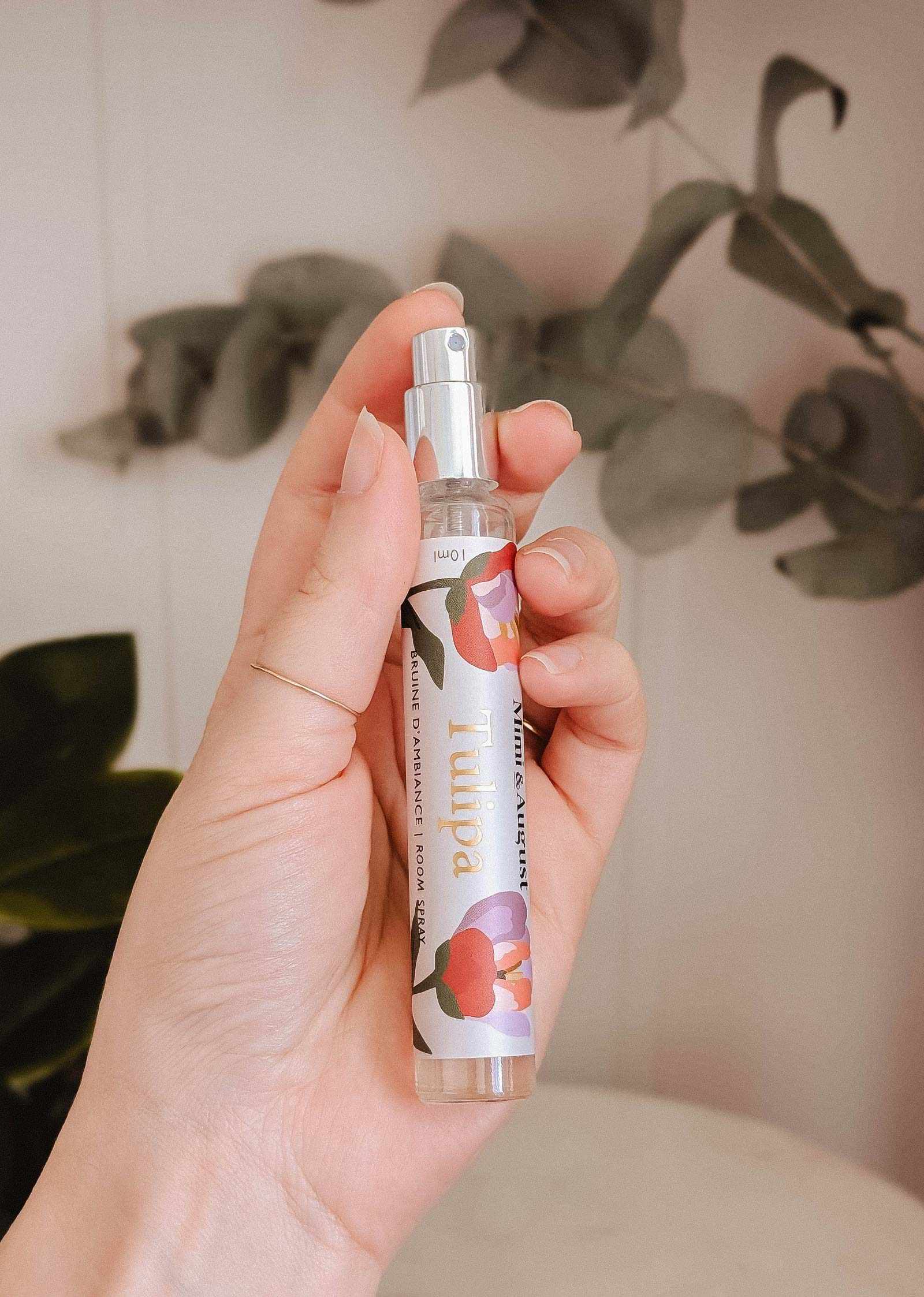 A hand holds a Mimi & August Room spray - Tulipa bottle with a floral design, evoking delicate floral notes, set against green leaves and a light-colored wall in the background.
