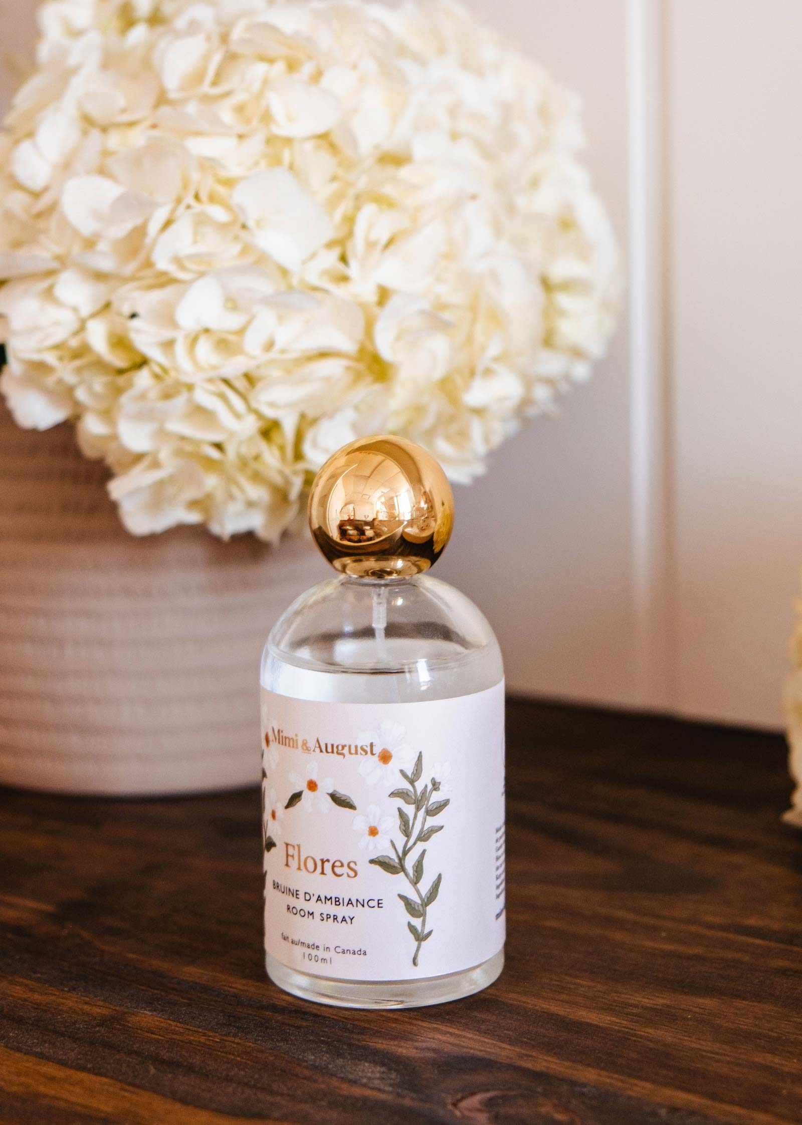 A clear bottle of Mimi & August Room spray Flores with a gold cap rests on a wooden surface in front of white hydrangeas, reflecting the floral essence of this room spray.