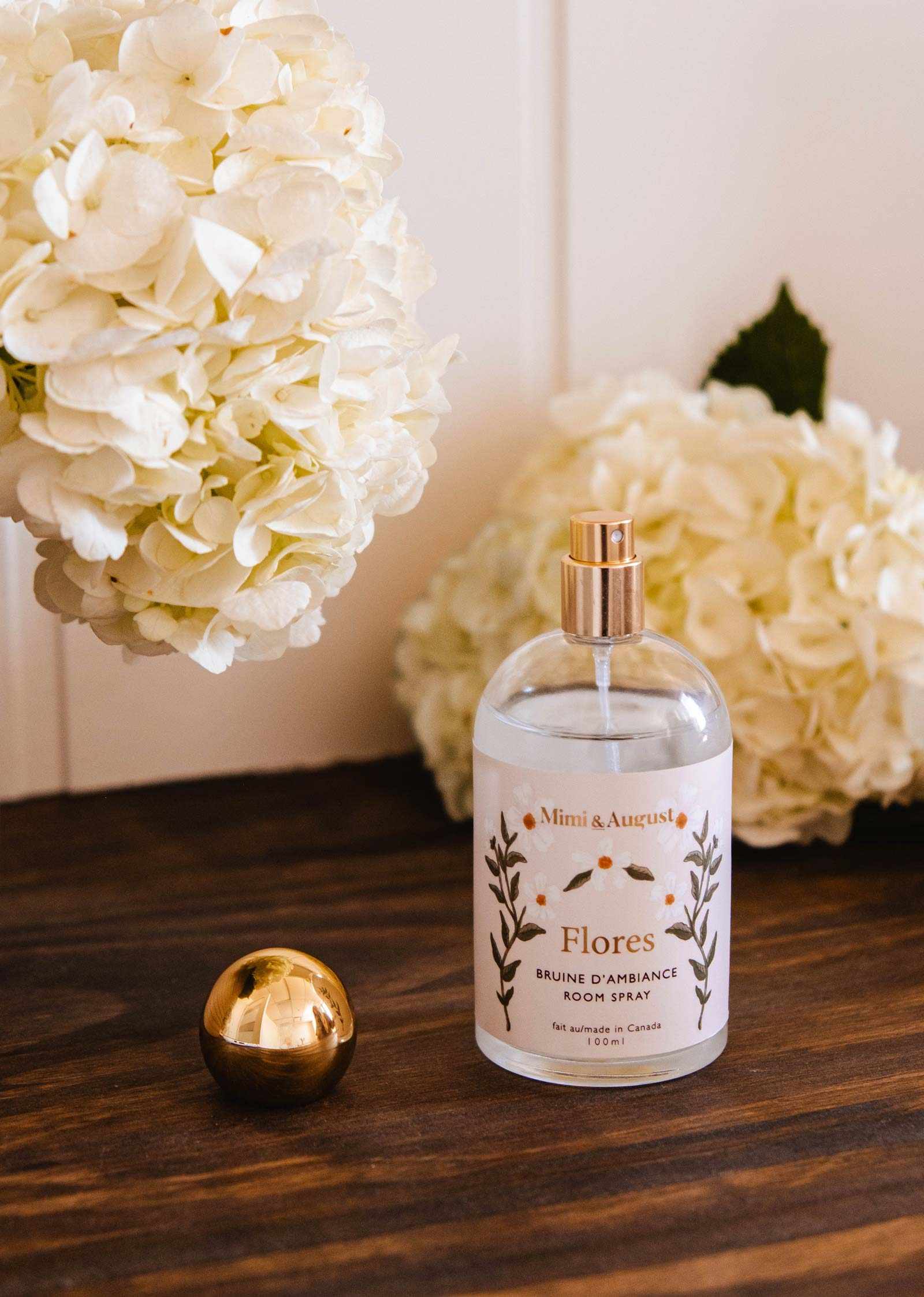 A clear bottle of Mimi & August Room Spray Flores with a gold cap rests on a wooden surface beside white hydrangeas, showcasing this elegant floral scent.
