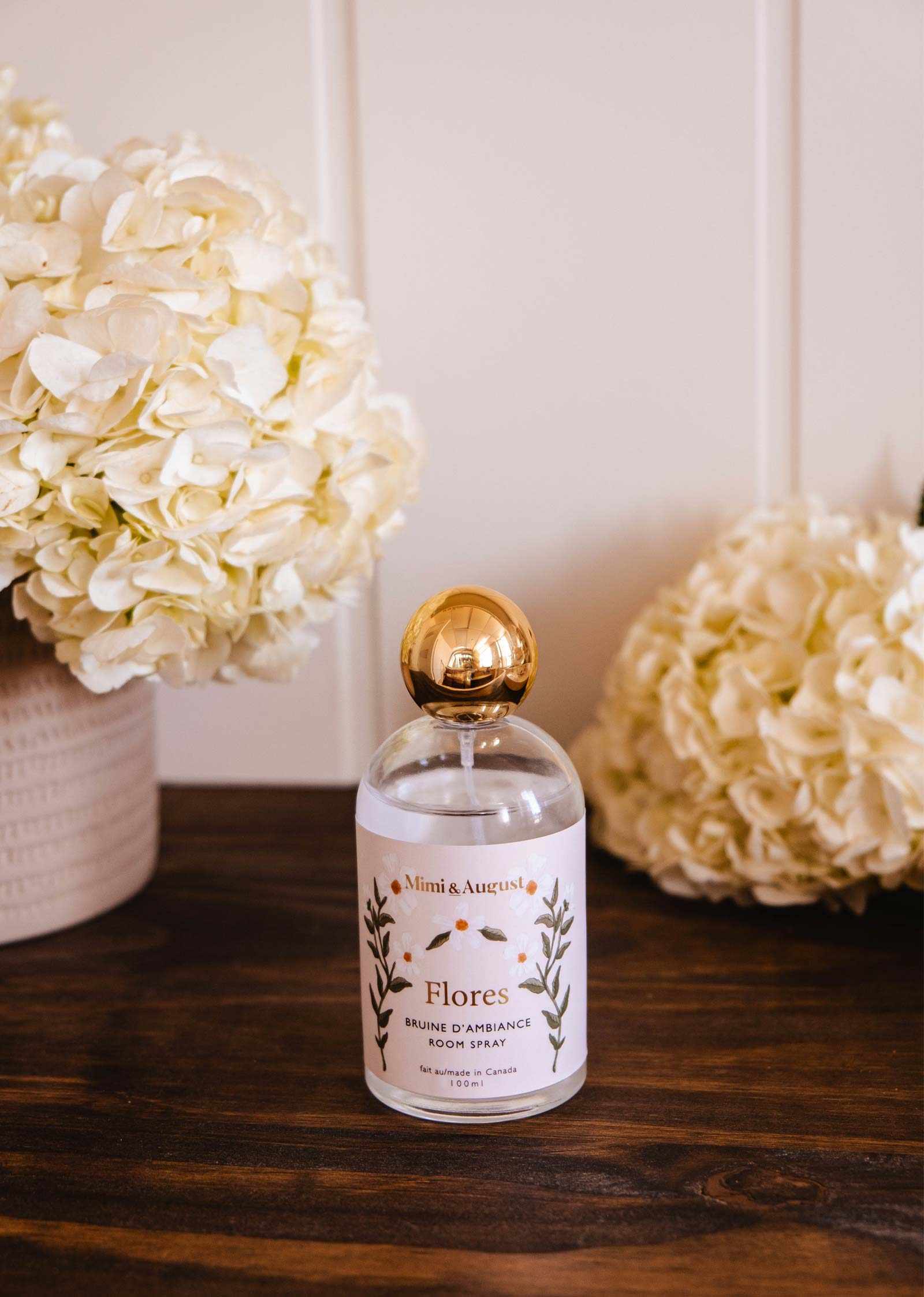 A bottle of Mimi & August Room Spray Flores with a gold cap rests on a wooden table, surrounded by white hydrangeas in vases, setting an elegant floral mood.