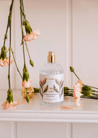 A Mimi & August Room spray - Oiseau du paradis, in a clear glass bottle with a floral label, sits on a shelf amidst light pink carnations, releasing delicate citrus and tropical fruit aromas.
