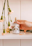 A hand holds a "Mimi & August" spray bottle beside pink flowers on a white surface, releasing a citrusy and tropical mist from the "Oiseau du paradis Room Spray," transforming any space into paradise.