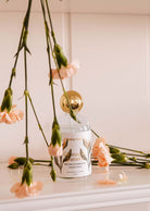 On a white surface, amid peach carnations, rests a Mimi & August Room Spray - Oiseau du paradis with a gold cap, emitting a citrusy and fruity tropical mist.