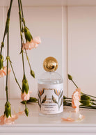 A Room spray - Oiseau du paradis bottle by Mimi & August, featuring a gold cap, elegantly sits on a shelf. Its fruity and tropical fragrance mingles with light pink blooms and green stems, enhancing the citrus notes for an exotic aroma reminiscent of paradise.