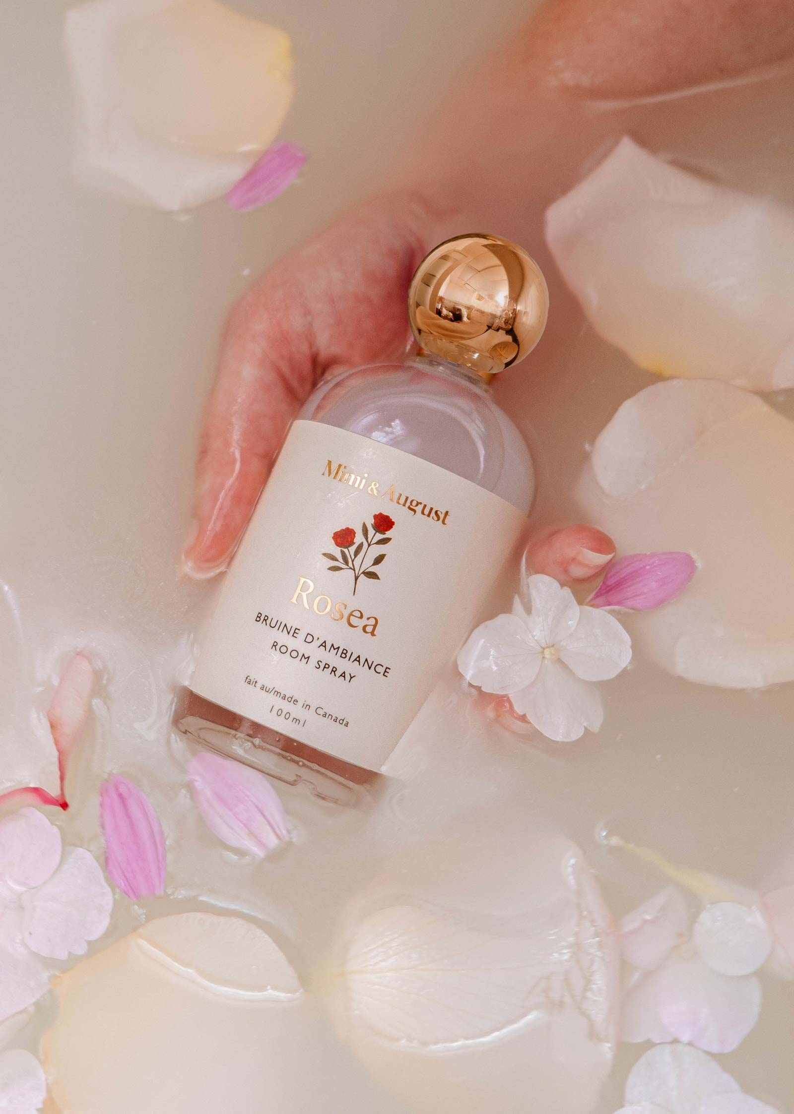 A hand holds a Mimi & August Room spray - Rosea bottle in a milky bath with rose petals and white flowers, showcasing this citrus floral fragrance as an elegant home scent option.