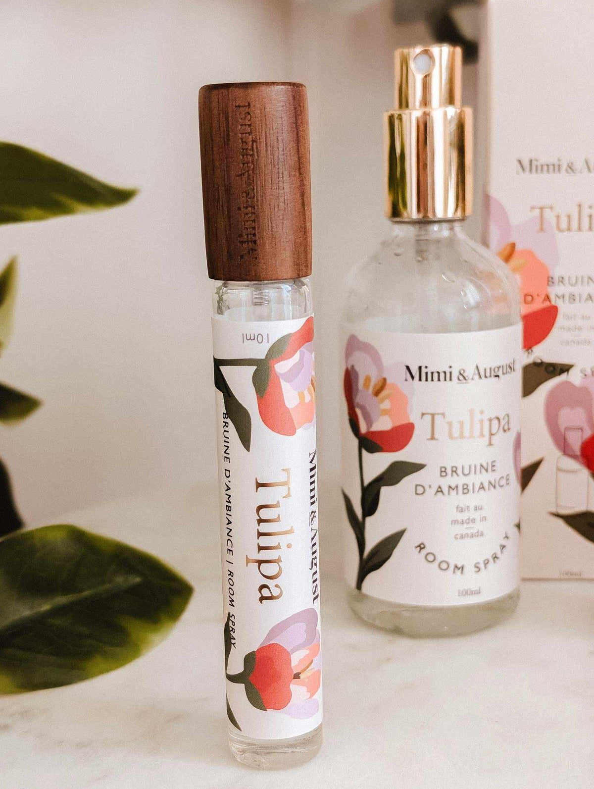 A glass Room spray Tulipa and a roller bottle, both by Mimi & August, are shown with floral designs and packaging in the background, suggesting a fresh wild berry scent.