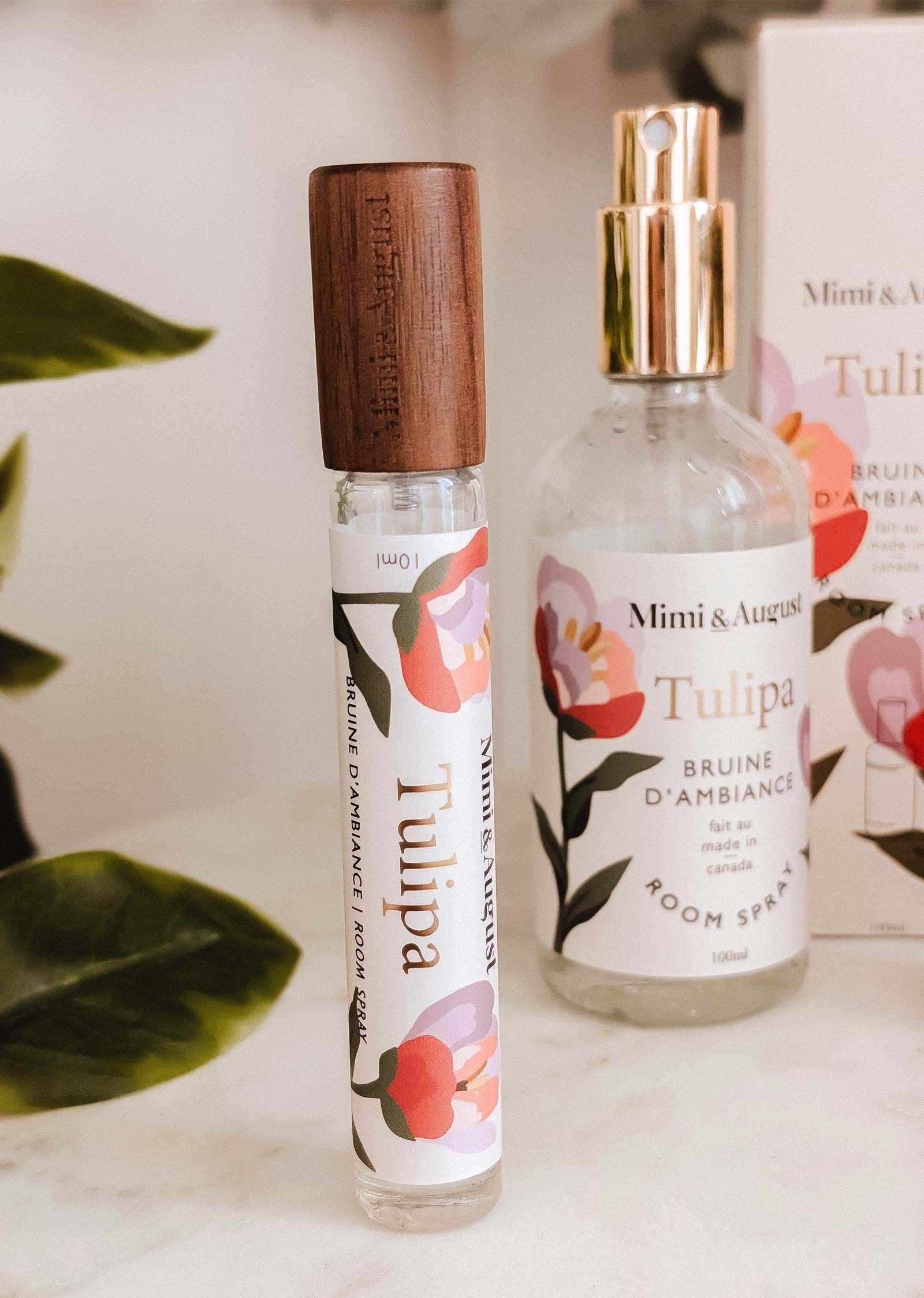 A glass Room spray Tulipa and a roller bottle, both by Mimi & August, are shown with floral designs and packaging in the background, suggesting a fresh wild berry scent.