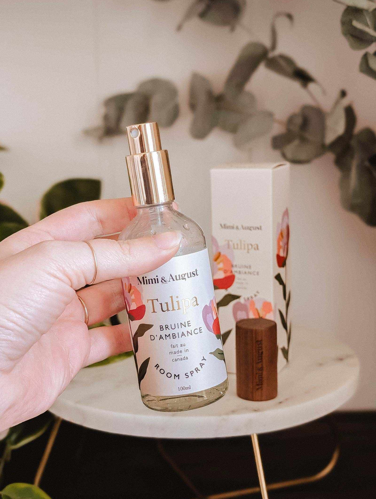 A hand holds a bottle of Room Spray Tulipa by Mimi & August in front of its packaging, displayed on a marble table with plants in the background.