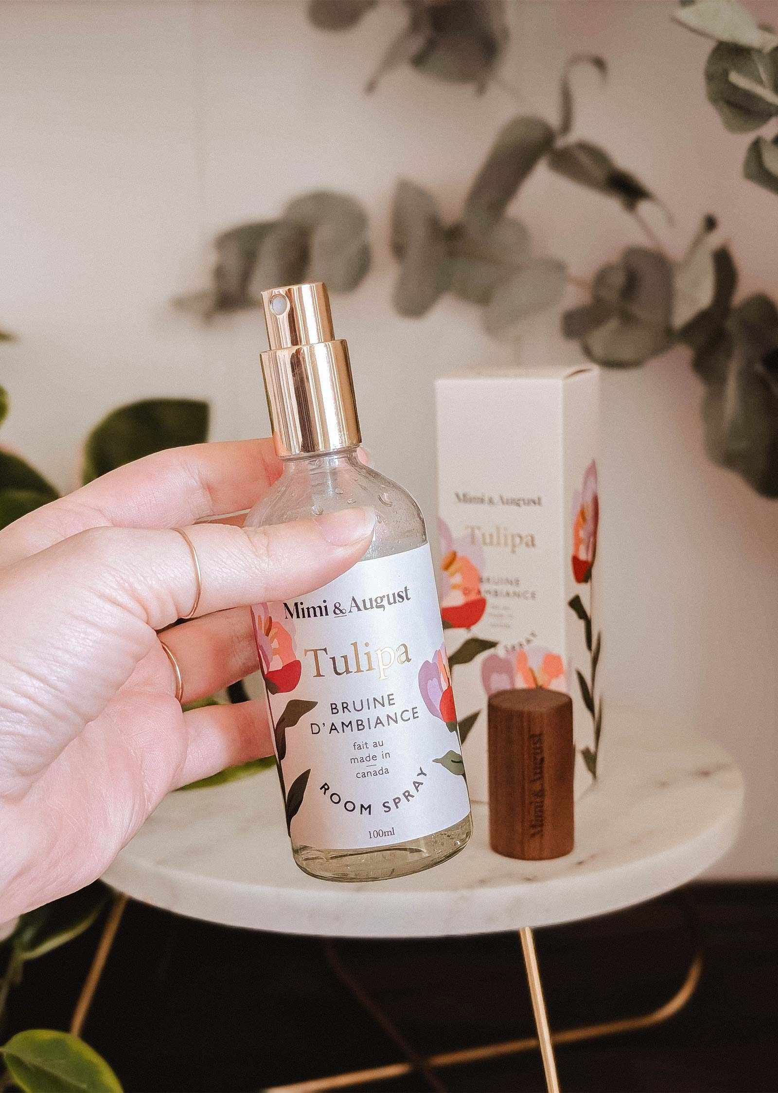 A hand holds a Mimi & August Room spray - Tulipa in front of its box, with green plants and a white table behind. This room spray features delicate floral notes for a refreshing atmosphere.