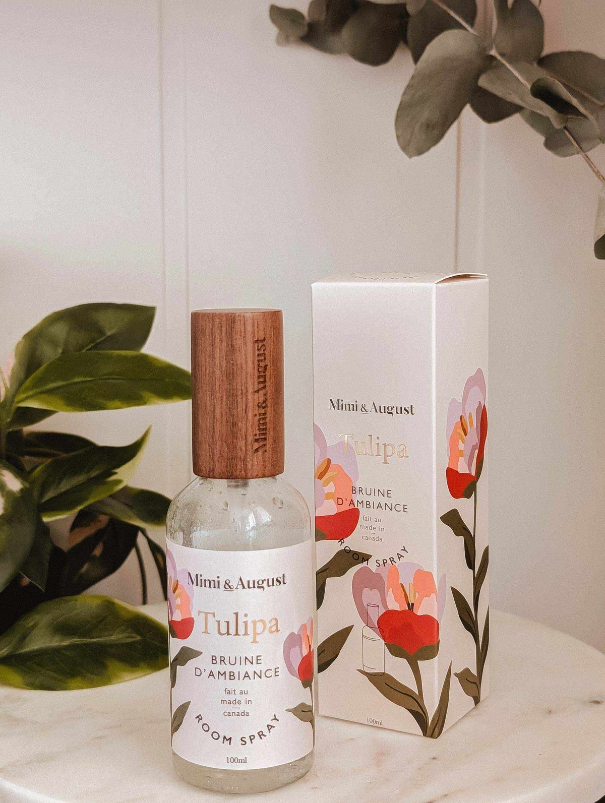 The Mimi & August Room Spray Tulipa with a wooden cap is displayed beside its matching floral box on a white surface, surrounded by green plants, highlighting this delightful floral scent.