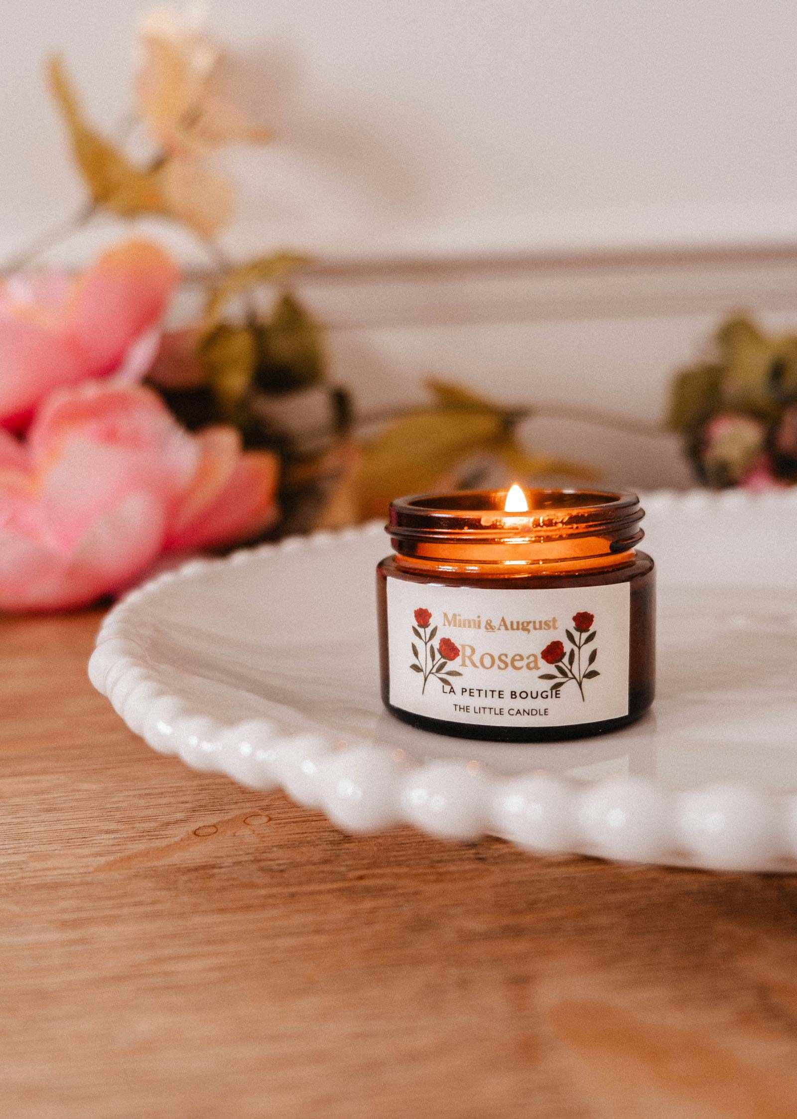 The Rosea – 2 oz Soy Candle by Mimi & August glows in a brown glass jar on a white plate, surrounded by pink flowers and greenery—a beautiful and fragrant soy candle.
