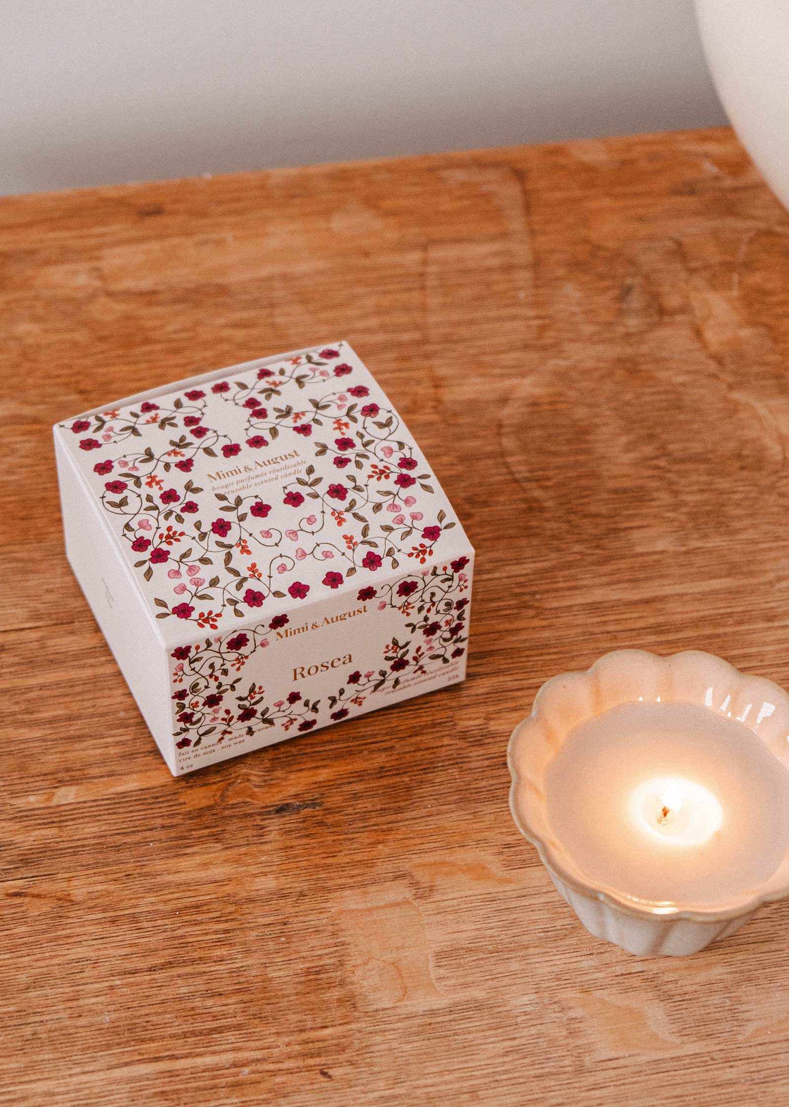 A floral-patterned Rosea - Reusable Candle by Mimi & August sits on a wooden surface in a white, scalloped-edge holder, radiating an uplifting scent.