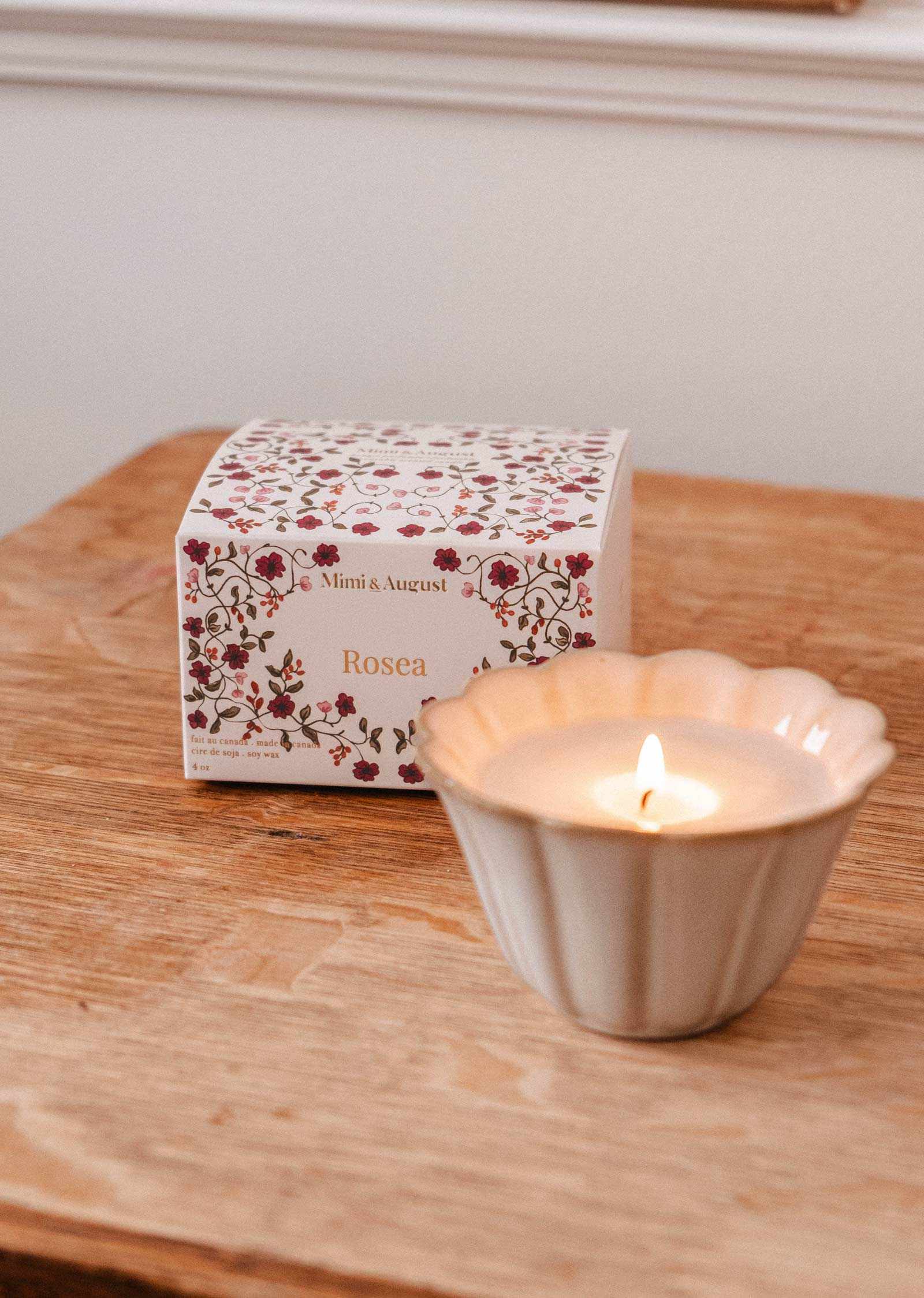 The hand-poured Rosea - Reusable Candle by Mimi & August glows in a scalloped holder on wood, topped with uplifting scent beside its floral-patterned box.