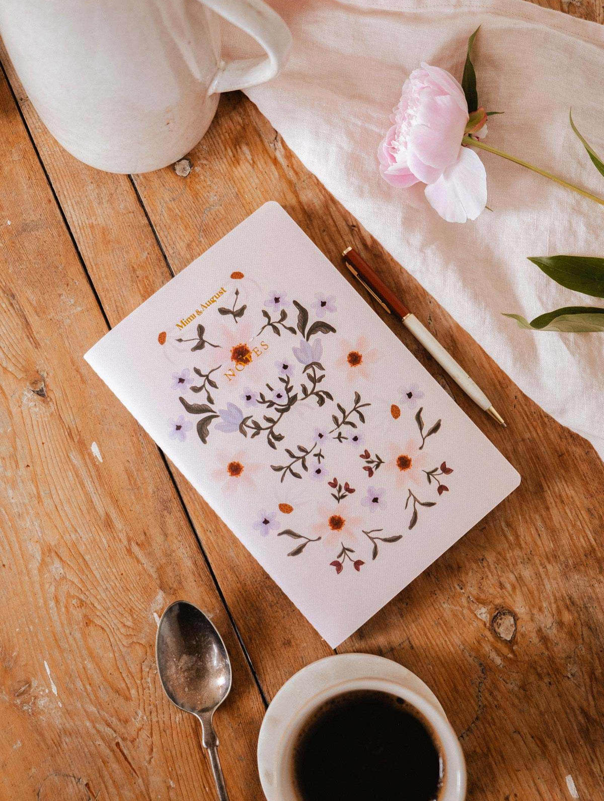 The Rosie notebook by Mimi & August, made with recycled paper, sits on a wooden table with a pen, spoon, coffee cup, white pitcher, and pink flower.