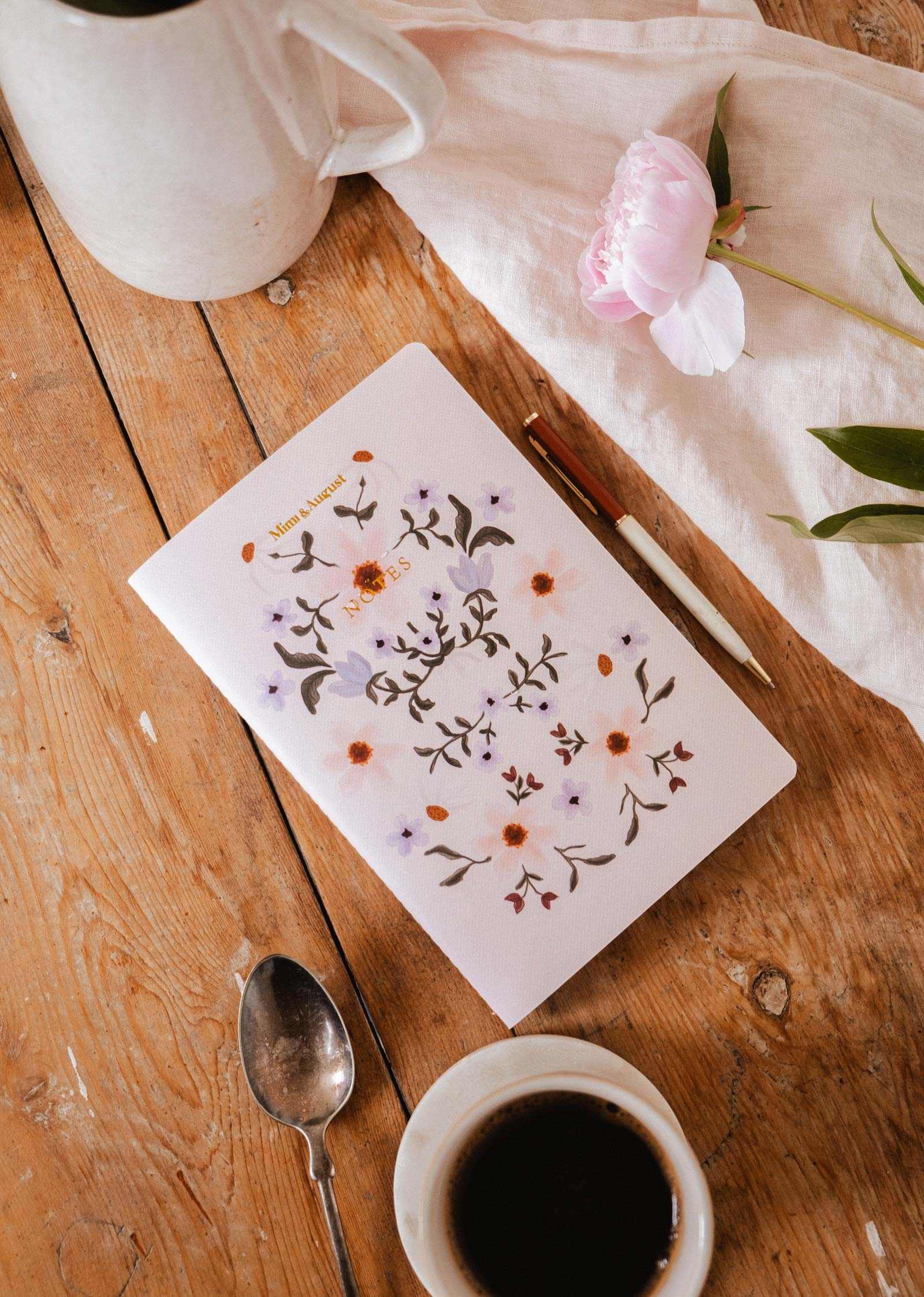 The Rosie notebook by Mimi & August, made with recycled paper, sits on a wooden table with a pen, spoon, coffee cup, white pitcher, and pink flower.