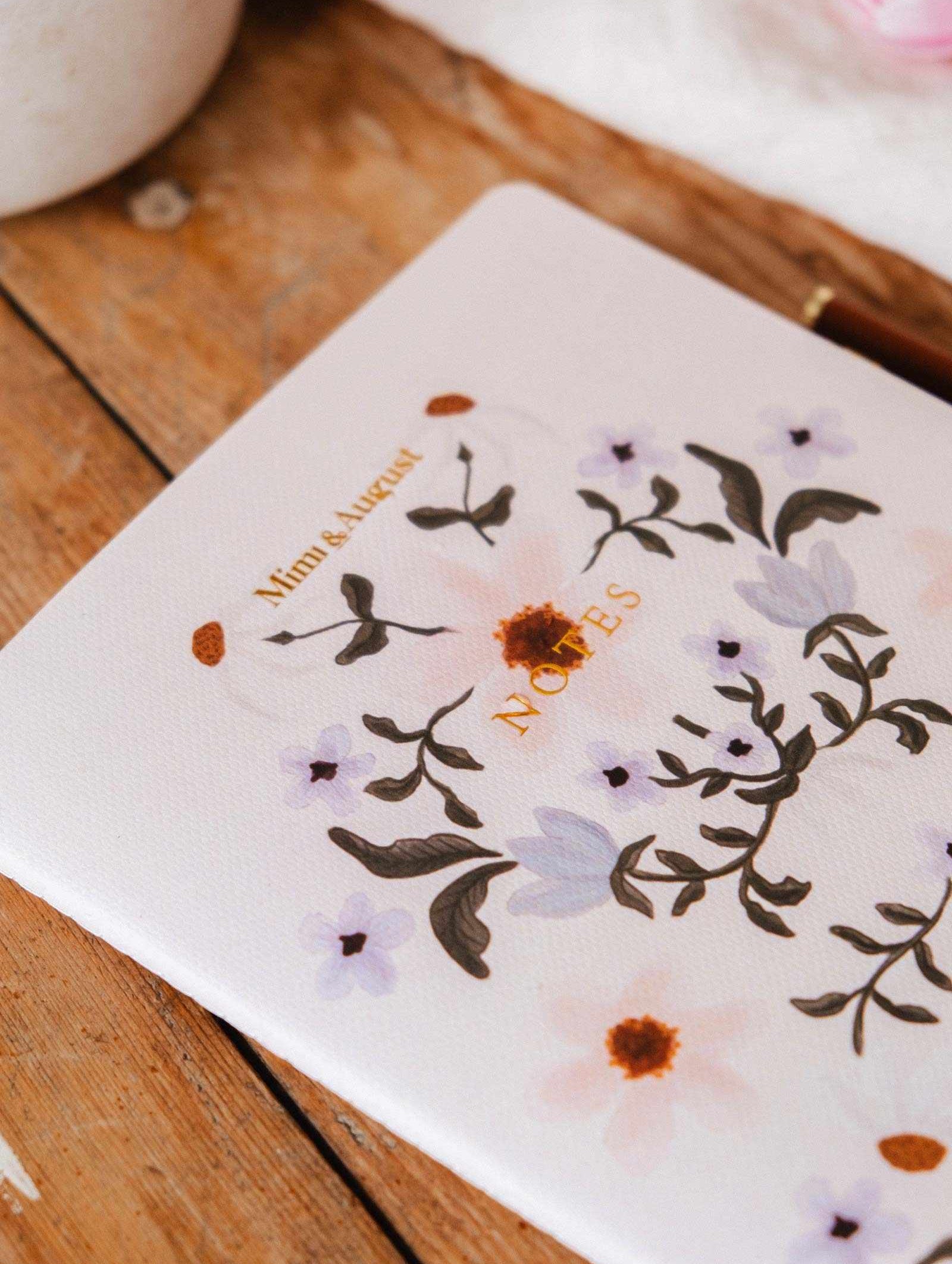 The Rosie Notebook by Mimi & August, made from recycled paper and featuring a charming floral design, rests on a wooden surface.