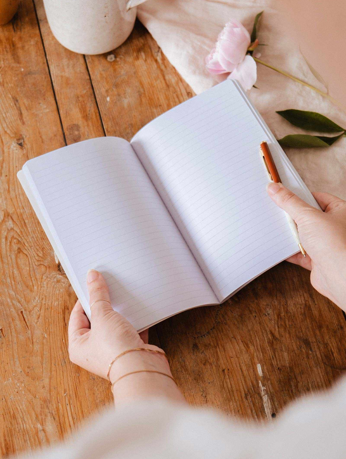 Someone holds an open Rosie - Notebook by Mimi & August with lined, floral pages and a pen on a wooden table; a pink flower and fabric are visible in the background.