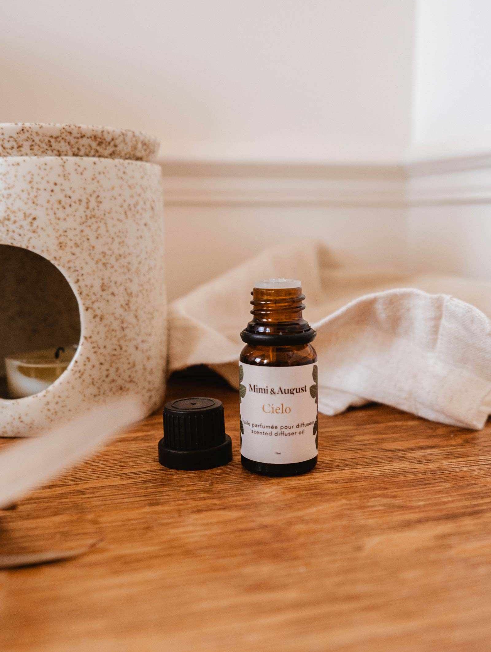 A brown bottle of Scented Oil - Cielo by Mimi & August with a black cap, positioned on a wooden surface beside a ceramic diffuser. The bottle's label reads "Mimi & August Cielo." The cap is off and resting next to it, allowing a calming scent to fill the air.