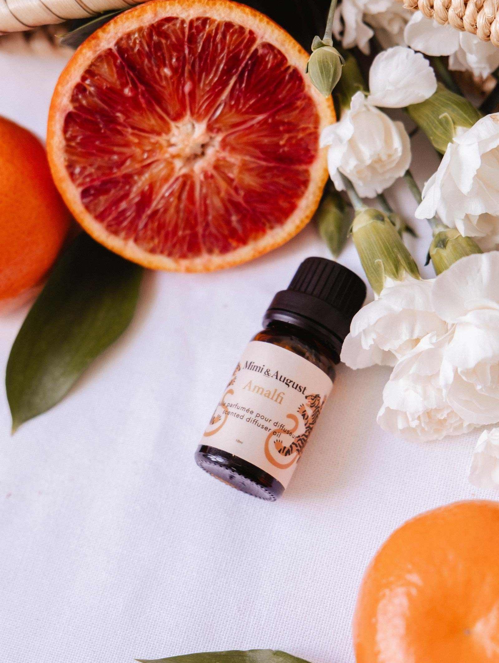 A small bottle of Mimi & August Scented Oil - Amalfi sits on a white surface with blood orange slices, whole oranges, white flowers, and green leaves—an inviting diffuser fragrance for relaxation and escape.
