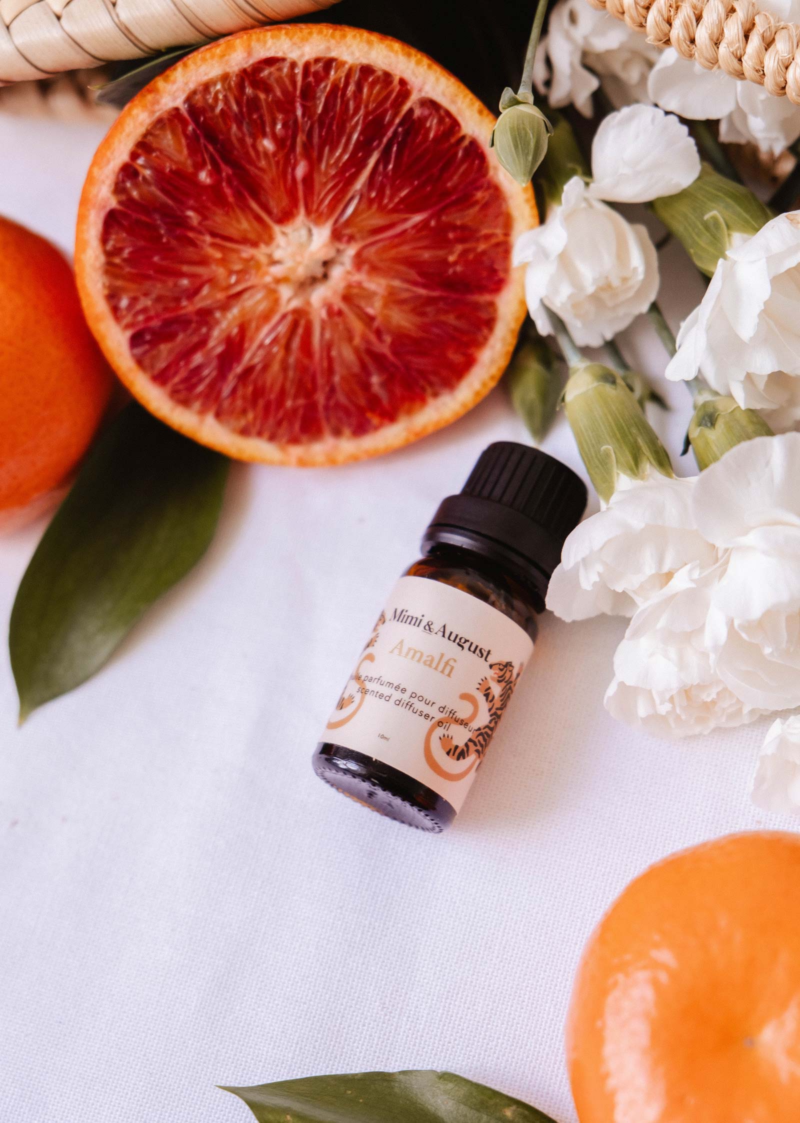 A small bottle of Mimi & August Scented Oil - Amalfi sits on a white surface with blood orange slices, whole oranges, white flowers, and green leaves—an inviting diffuser fragrance for relaxation and escape.