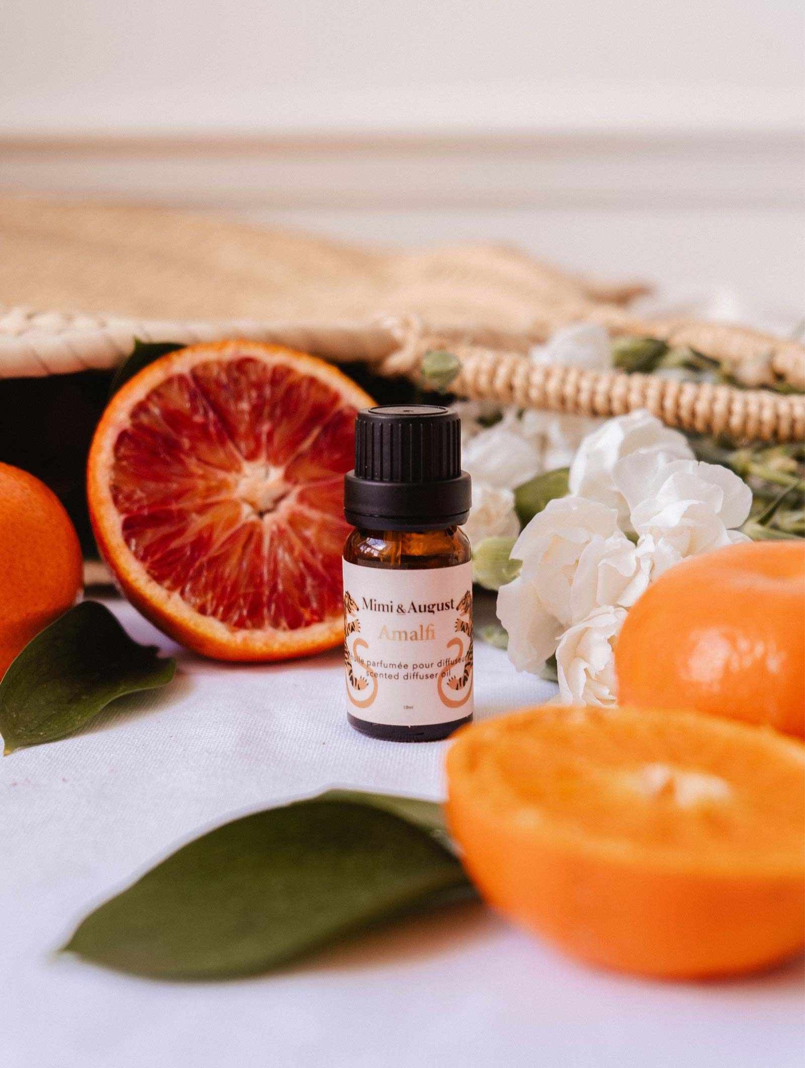 A small Mimi & August Scented Oil - Amalfi bottle sits on a white surface, surrounded by sliced oranges, a woven bag, green leaves, and white flowers—a picturesque scene for relaxation.