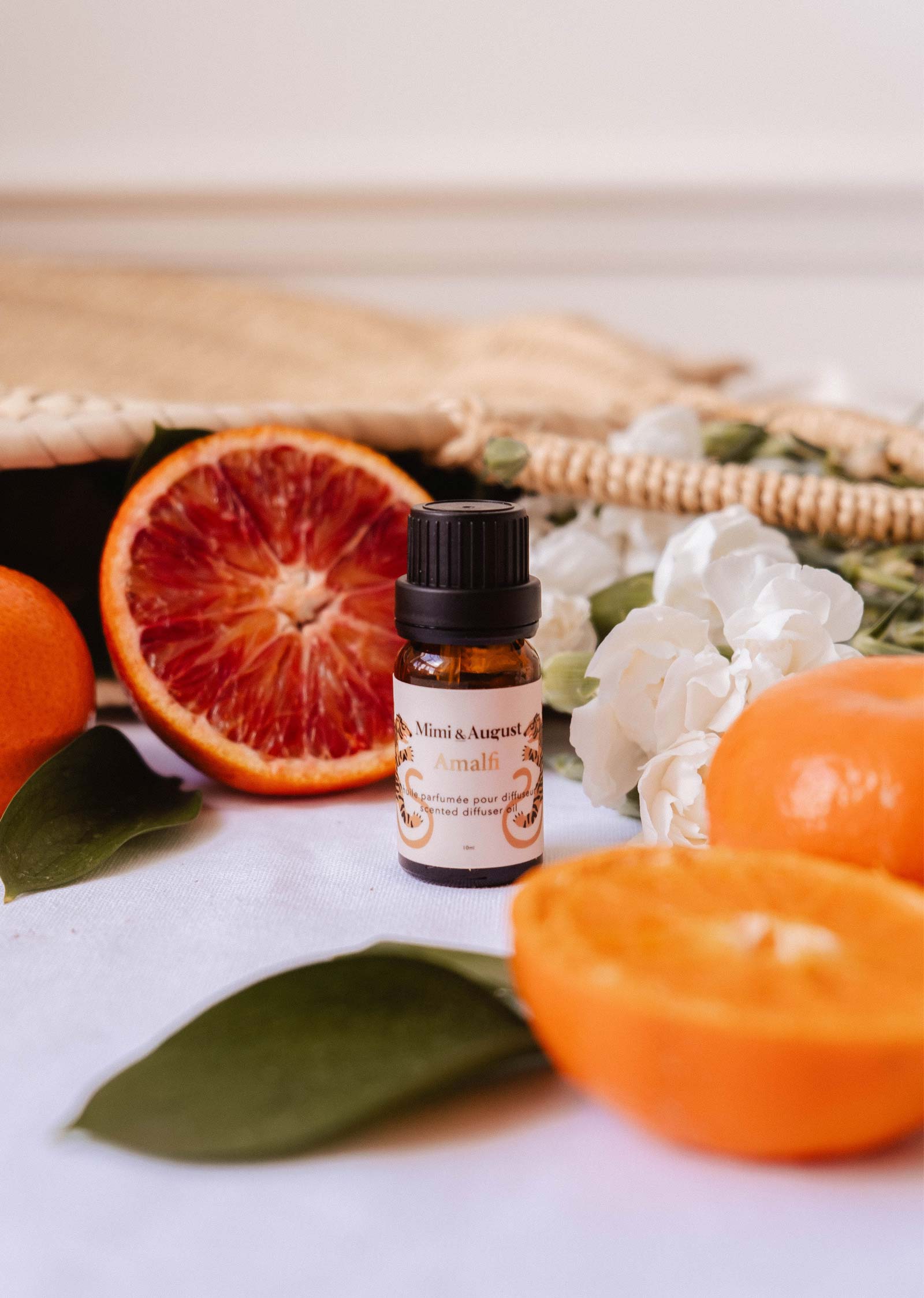 A small Mimi & August Scented Oil - Amalfi bottle sits on a white surface, surrounded by sliced oranges, a woven bag, green leaves, and white flowers—a picturesque scene for relaxation.