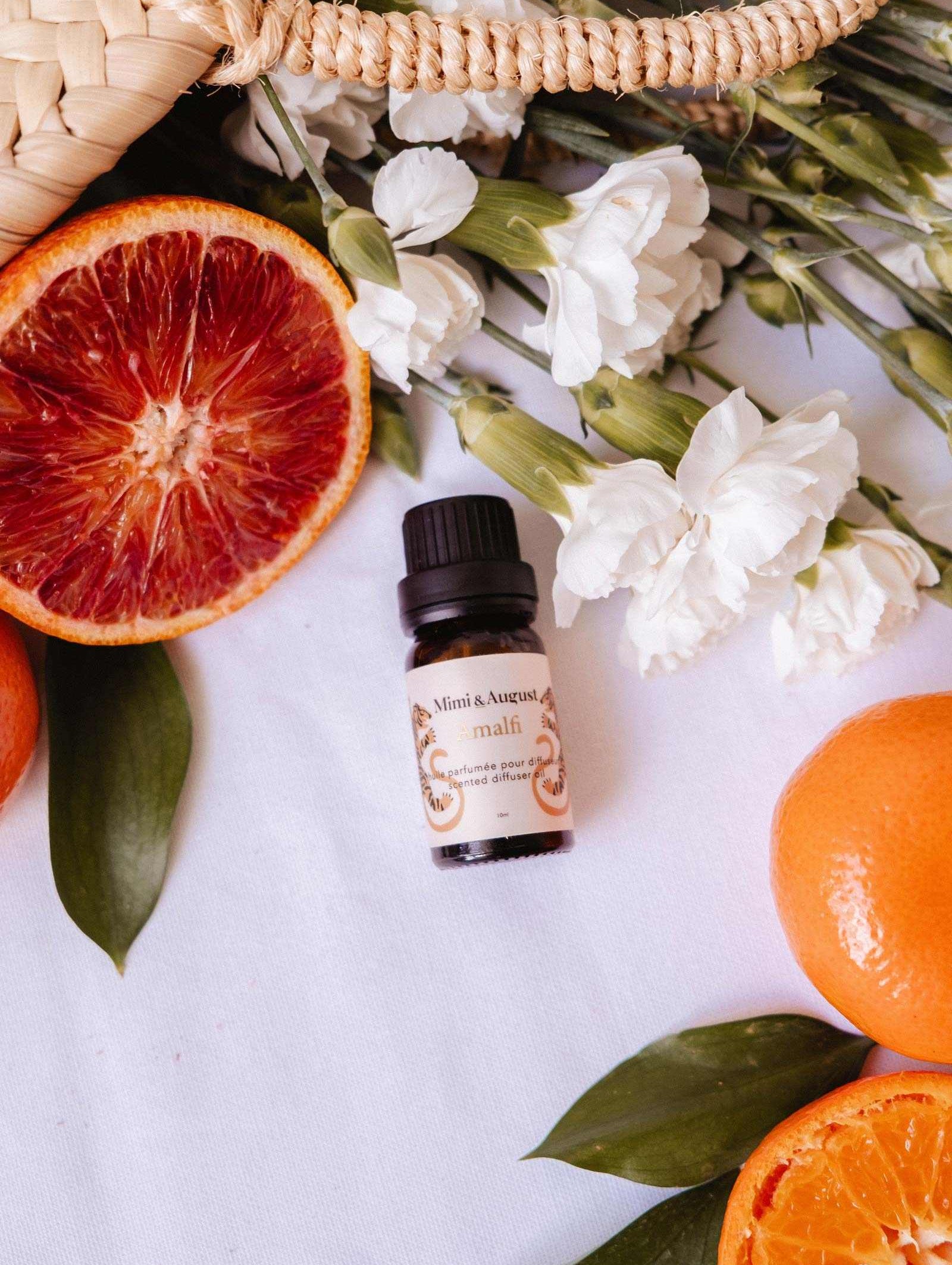Mimi & August Scented Oil - Amalfi is displayed on a white surface with white flowers, blood orange halves, and whole mandarins, creating an inviting sense of relaxation and escape.