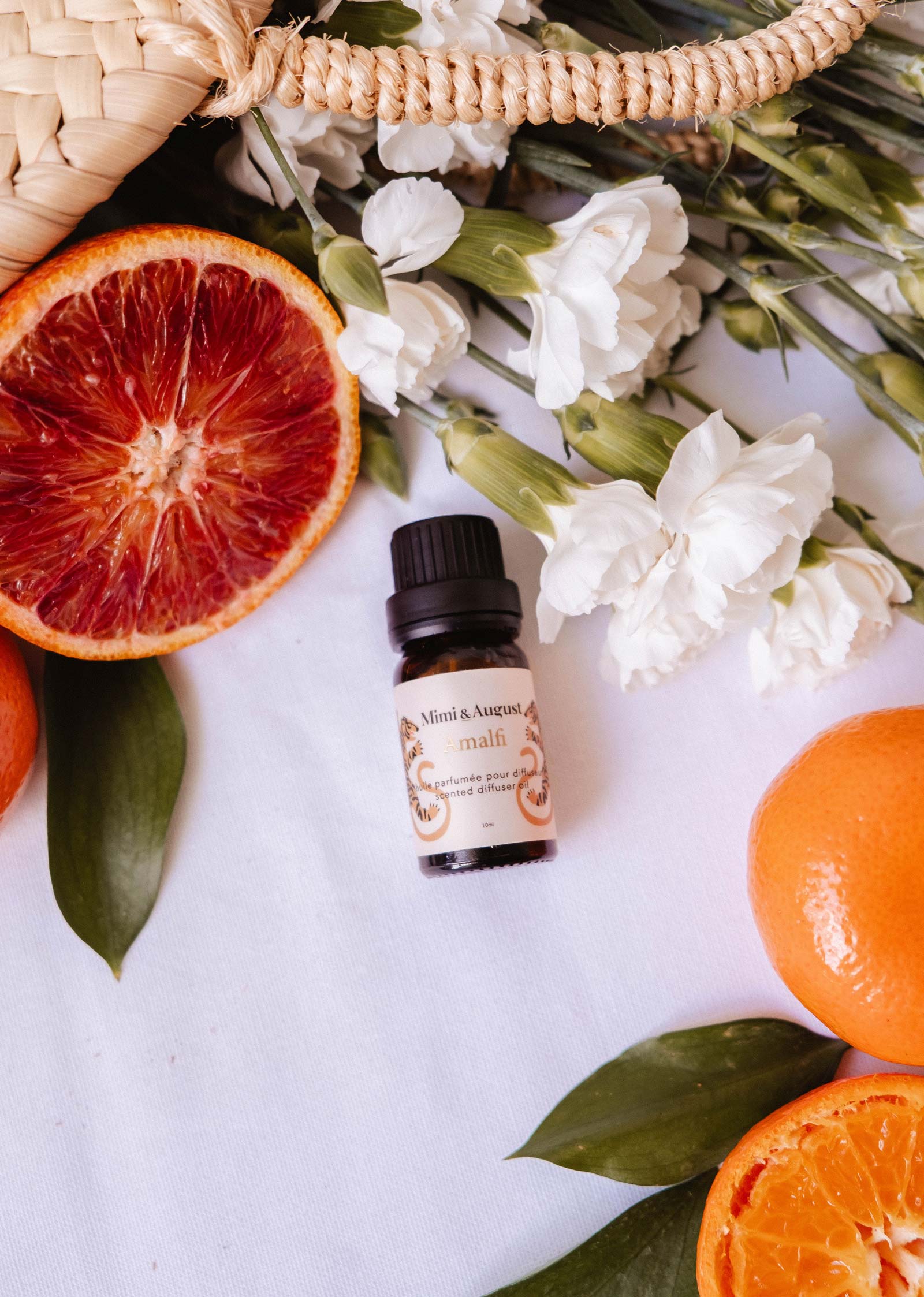 Mimi & August Scented Oil - Amalfi is displayed on a white surface with white flowers, blood orange halves, and whole mandarins, creating an inviting sense of relaxation and escape.