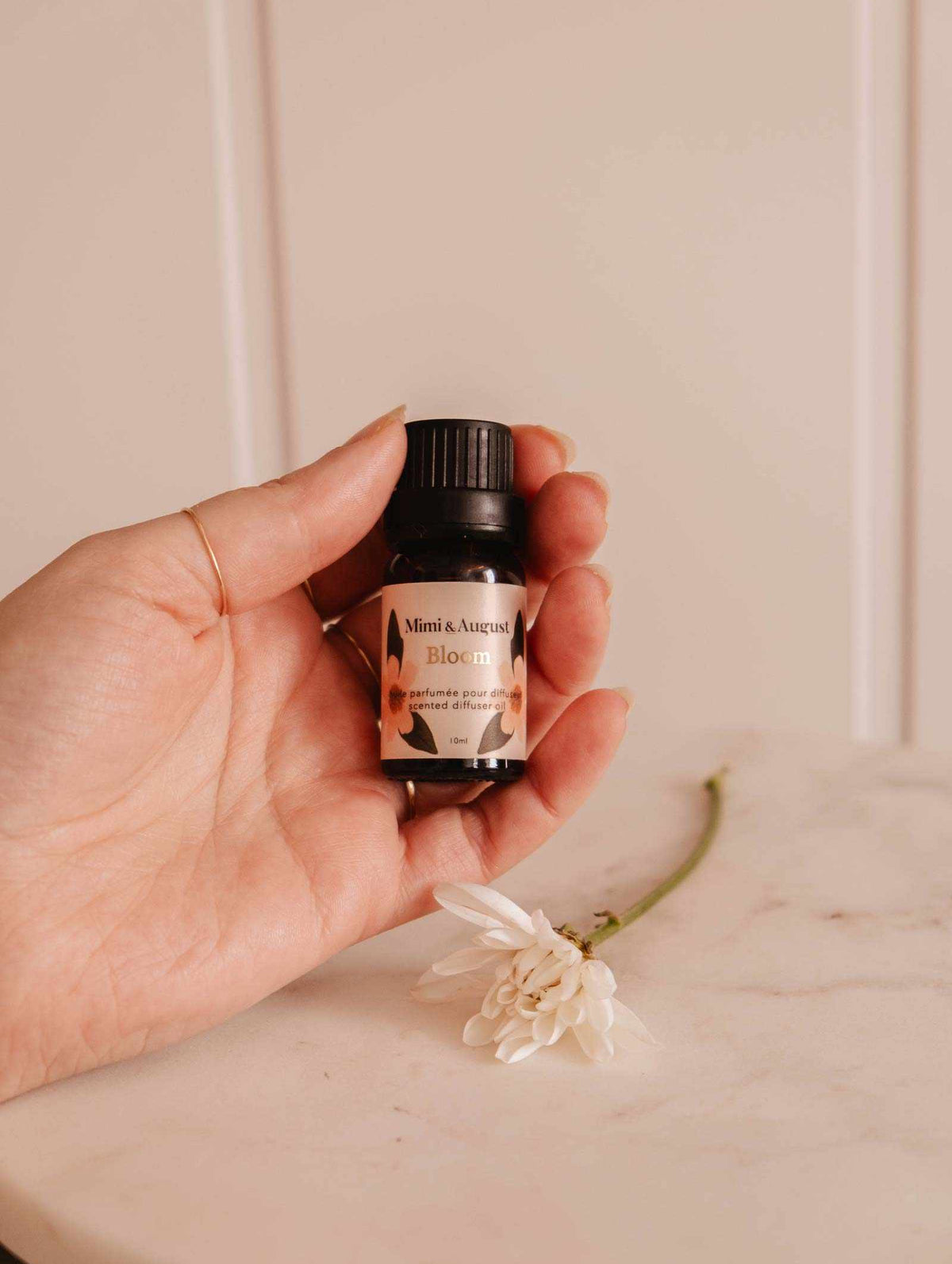 A hand holds a small bottle labeled "Mimi & August Scented Oil - Bloom," exuding a refreshing fragrance with a white flower on a marble surface.
