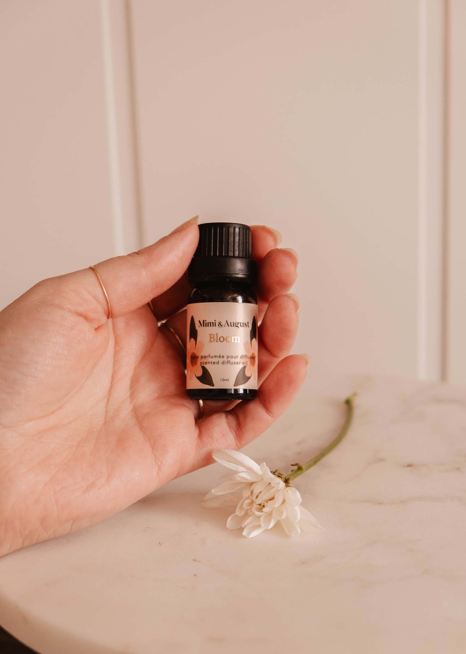 A hand holds a small bottle labeled "Mimi & August Scented Oil - Bloom," exuding a refreshing fragrance with a white flower on a marble surface.