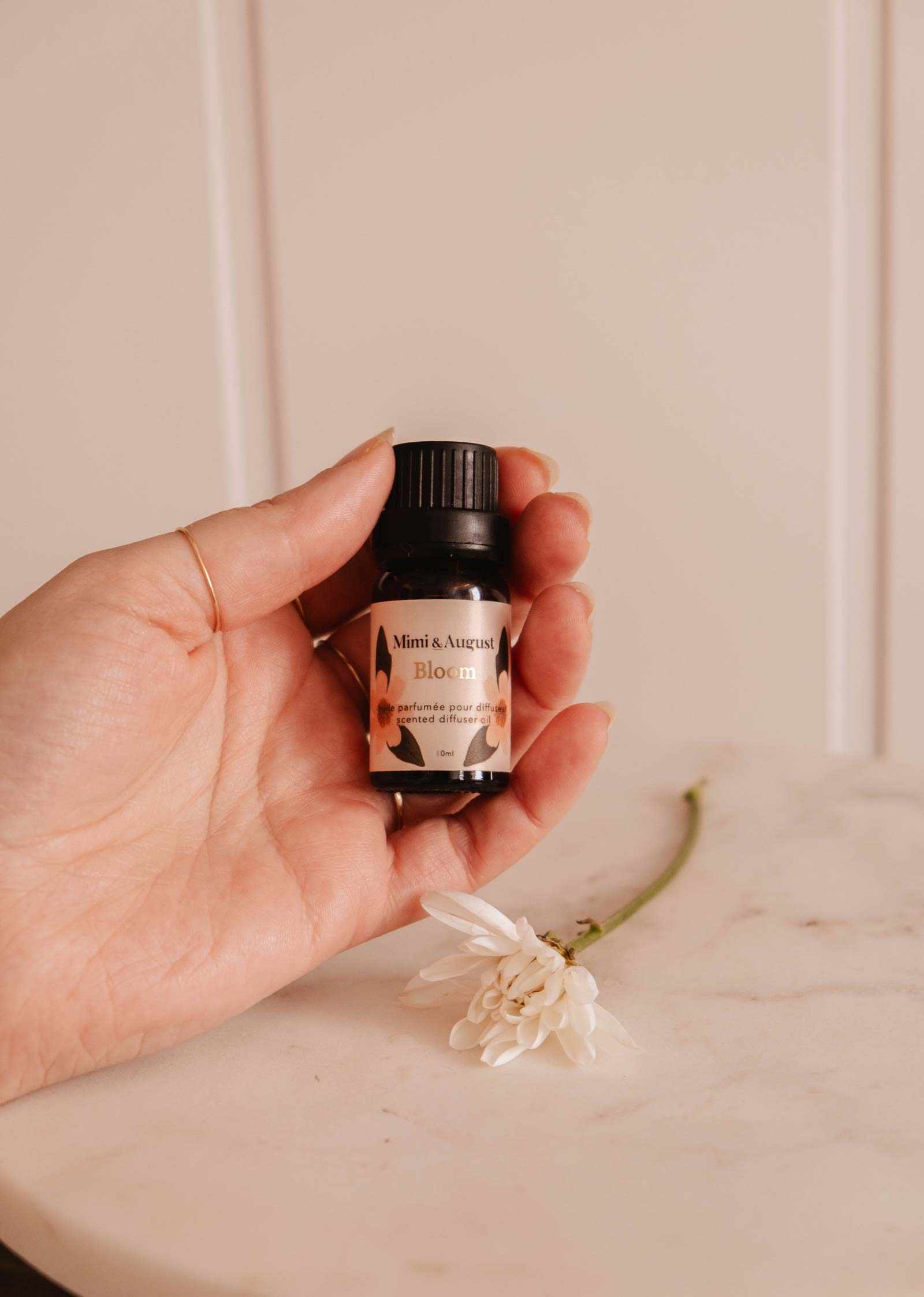 Une main tient une petite bouteille étiquetée « Mimi & August Huile parfumée – Bloom », dégageant une fragrance rafraîchissante avec une fleur blanche sur une surface en marbre.